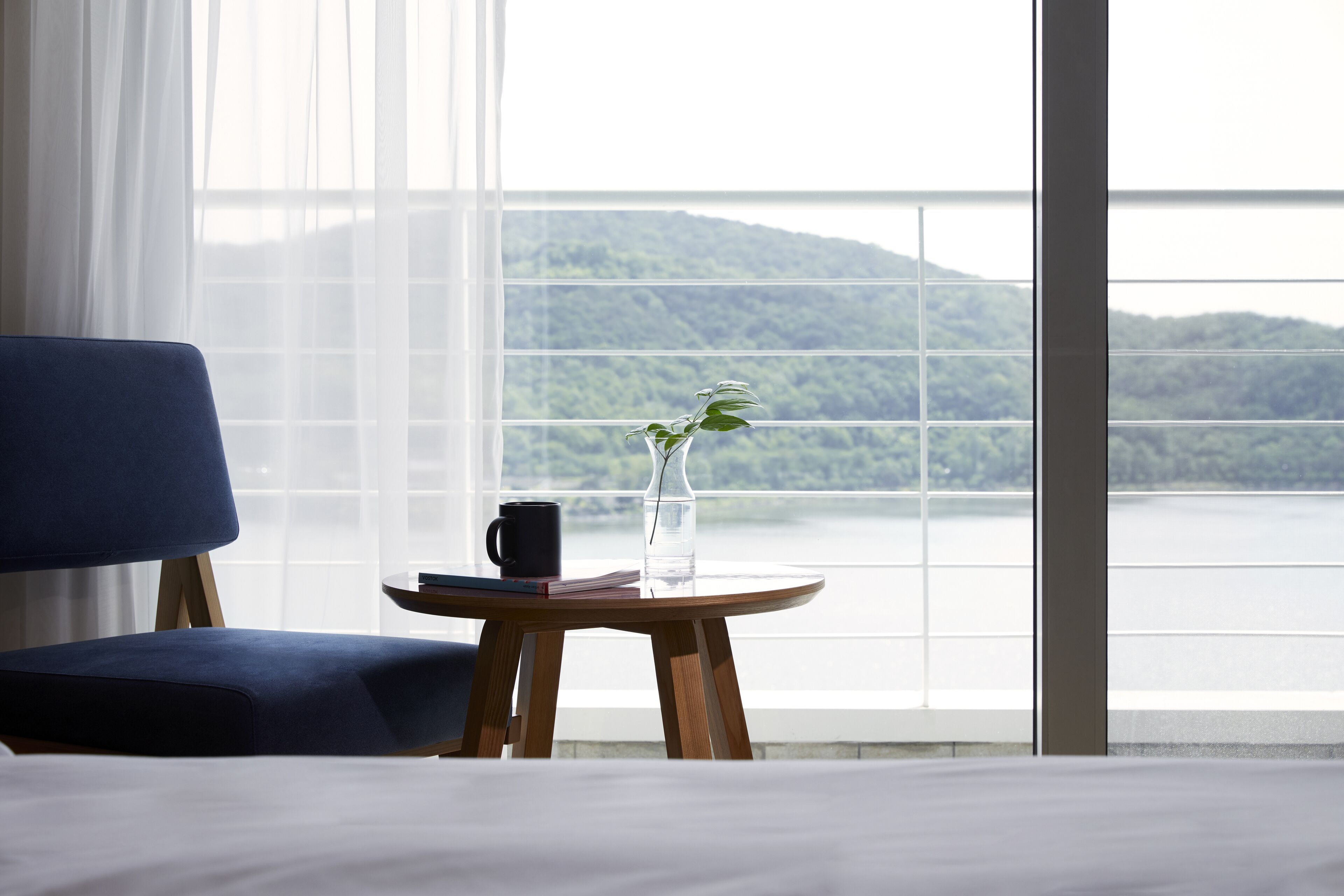 Una habitación con una cama, una silla azul, una pequeña mesa redonda de madera con un jarrón y una taza negra, y un ventanal con vistas a un lago y montañas.
