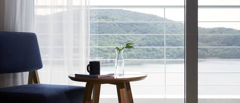 Una habitación con una cama, una silla azul, una pequeña mesa redonda de madera con un jarrón y una taza negra, y un ventanal con vistas a un lago y montañas.