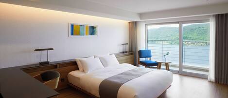 Une chambre dâhĂŽtel comprenant un lit, un bureau, une chaise, une petite table avec une plante et une grande fenĂȘtre donnant sur un plan dâeau et des collines.