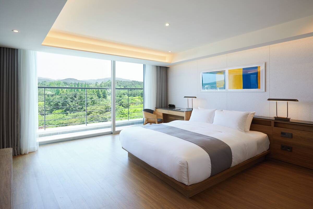 A modern hotel room with a large bed, a desk, and a view of a green landscape.