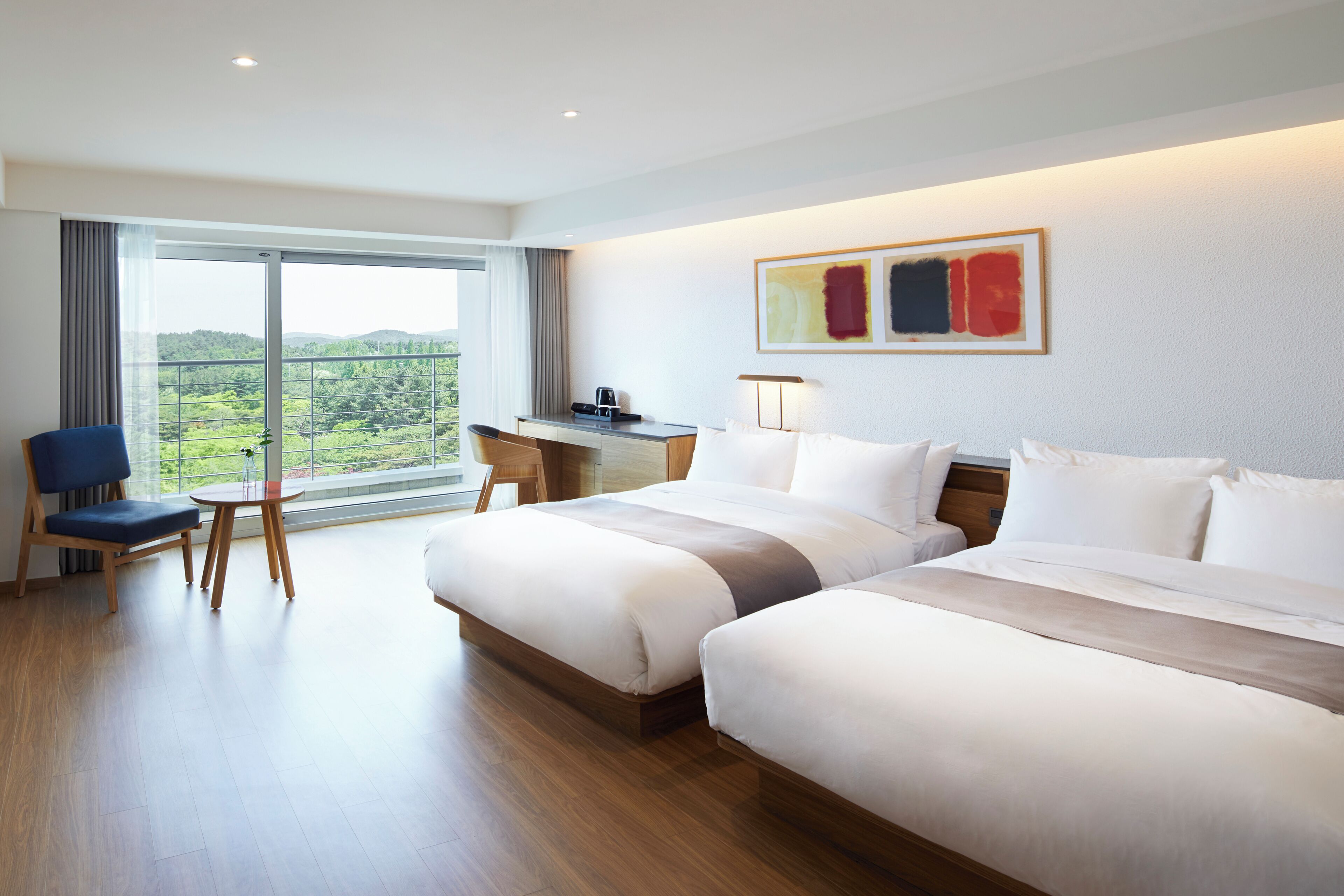 Ein Hotelzimmer mit zwei Betten, einem Schreibtisch, einem Sessel und einem großen Fenster mit Blick auf Grünflächen.