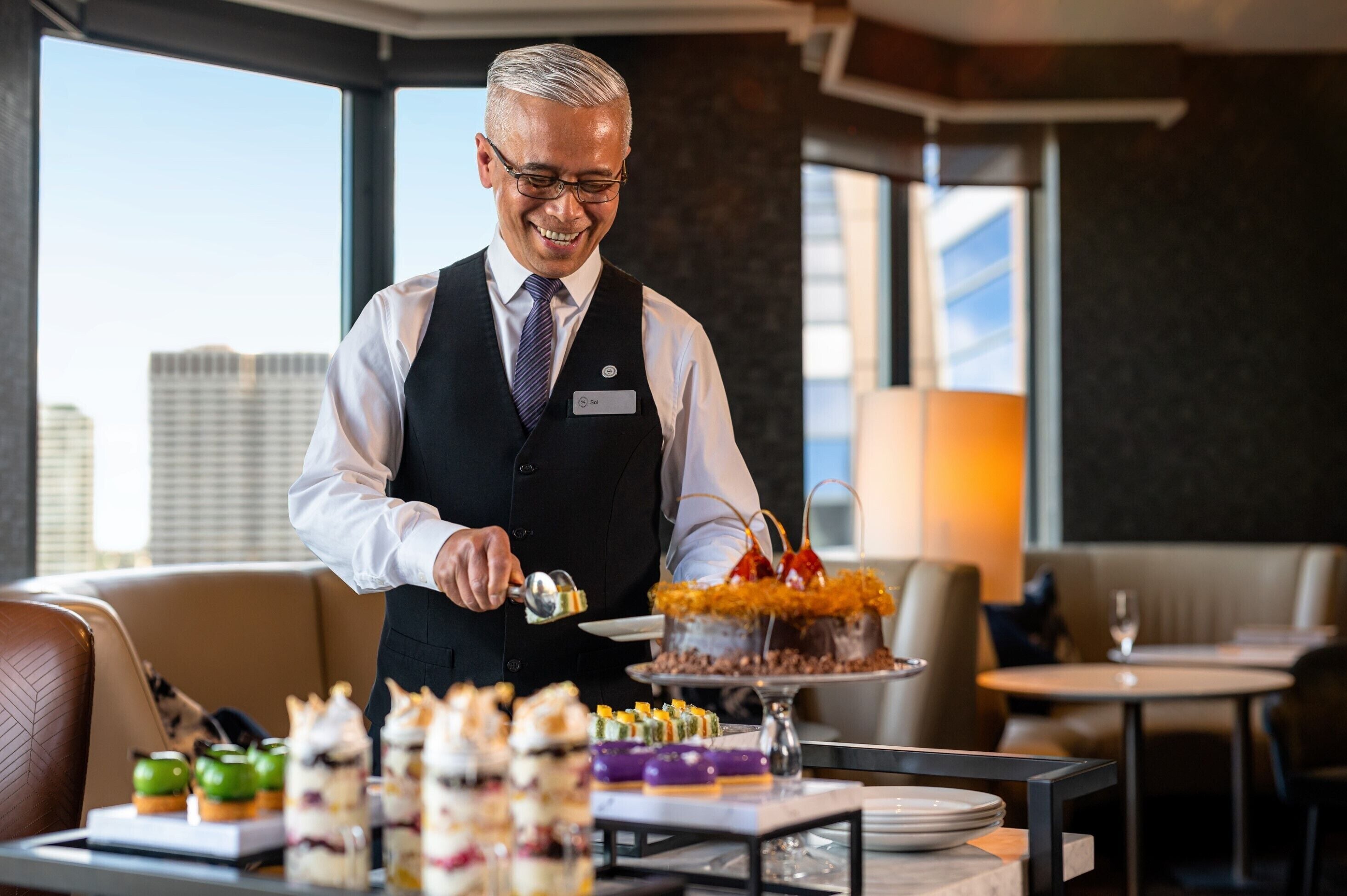A waiter serving a dessert in a modern restaurant with a city view.