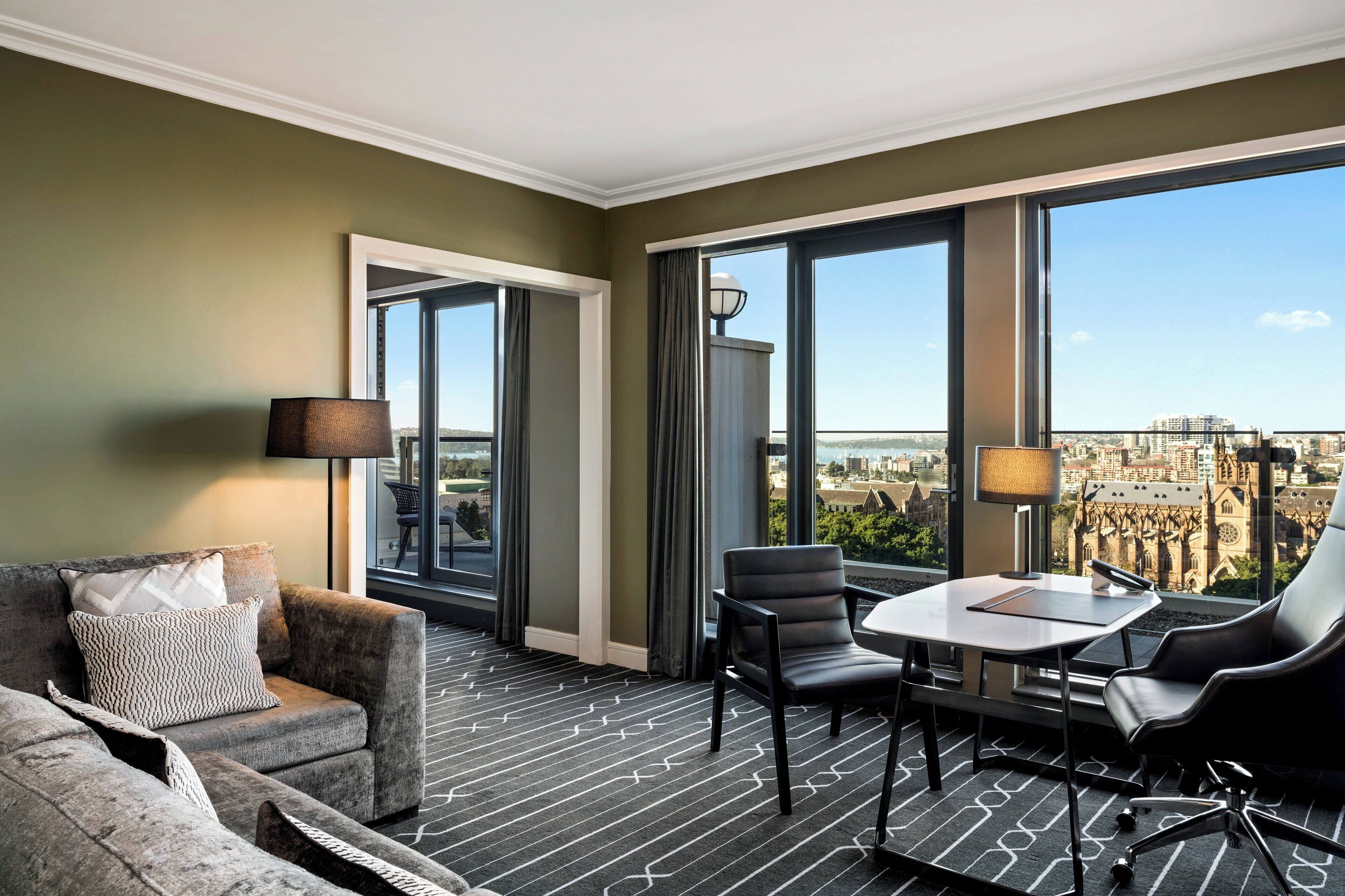 A modern hotel room with a large window offering a city view, a sofa, a table with chairs, and a lamp.