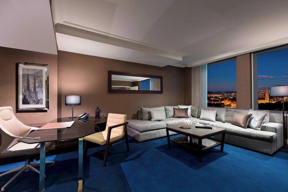 Een moderne hotelkamer met een groot raam en uitzicht over de stad, een bank, een bijzettafeltje, een bureau met stoel en een ingelijste foto aan de muur.