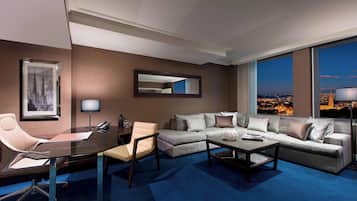 A modern hotel room with a large window offering a city view, a sofa, a coffee table, a desk with a chair, and a framed picture on the wall.