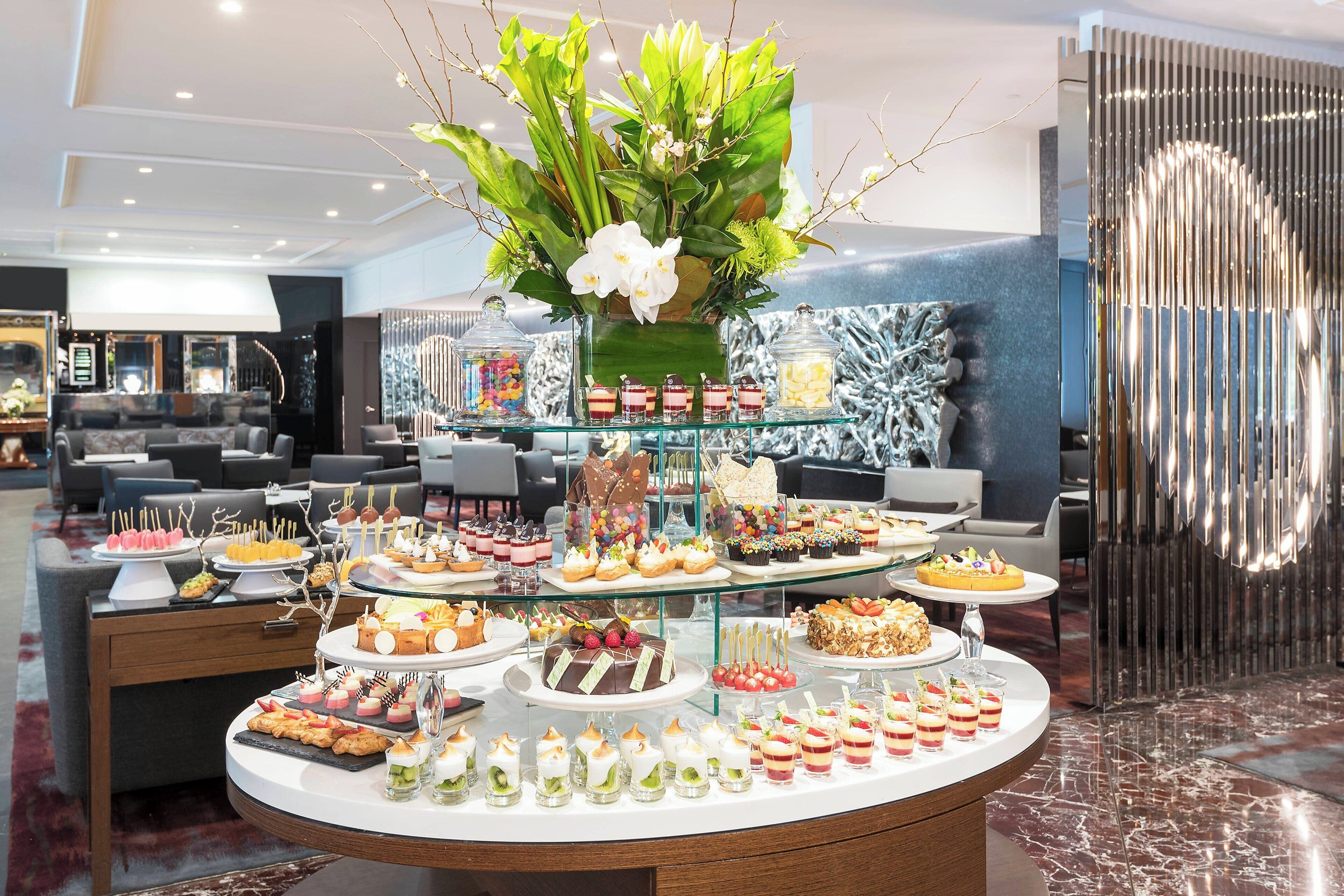 Un bufé con diversos postres y aperitivos, un centro de mesa floral y un interior moderno con zonas de estar.