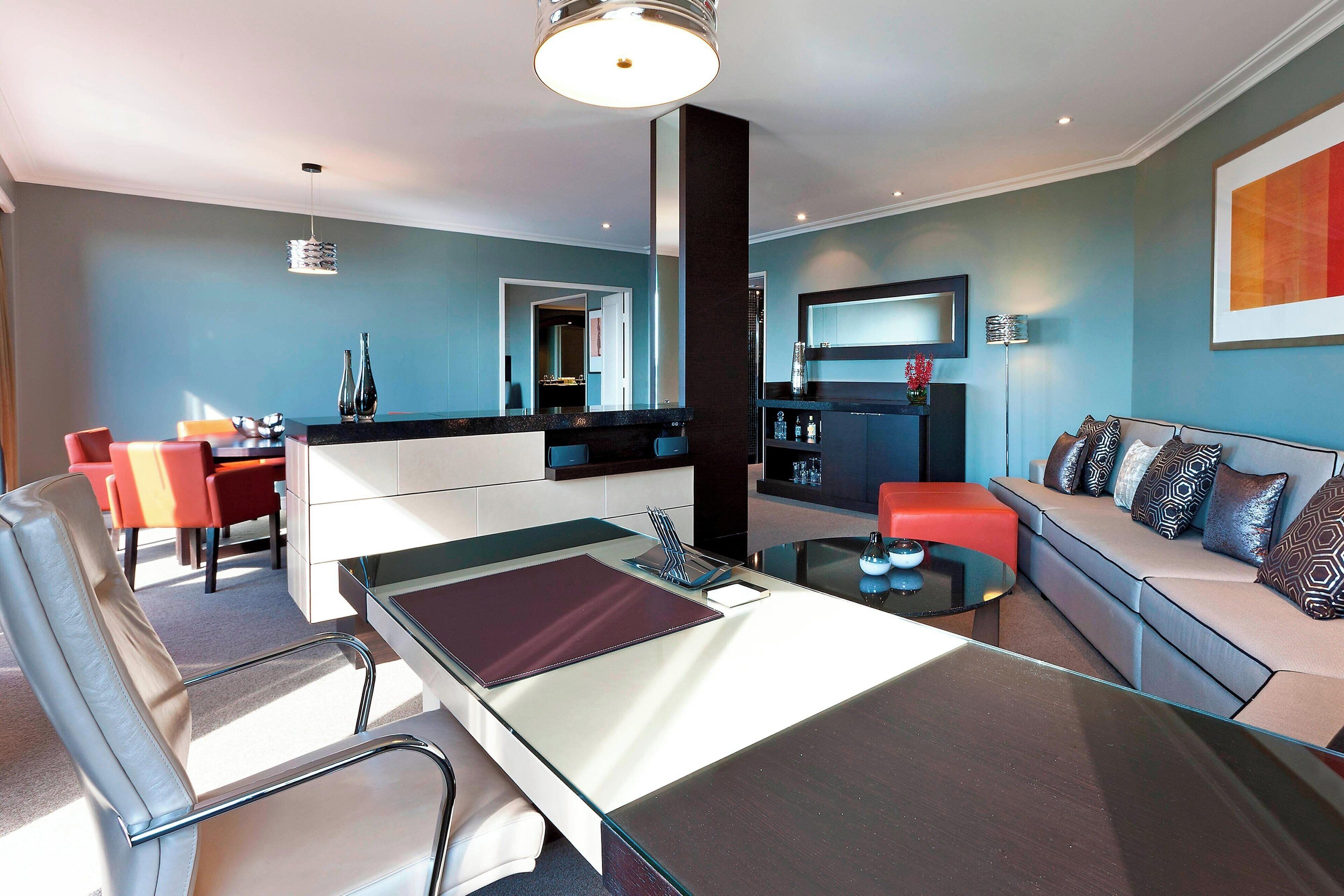 A modern living room with a glass dining table, a sofa with patterned cushions, and a kitchen area in the background.