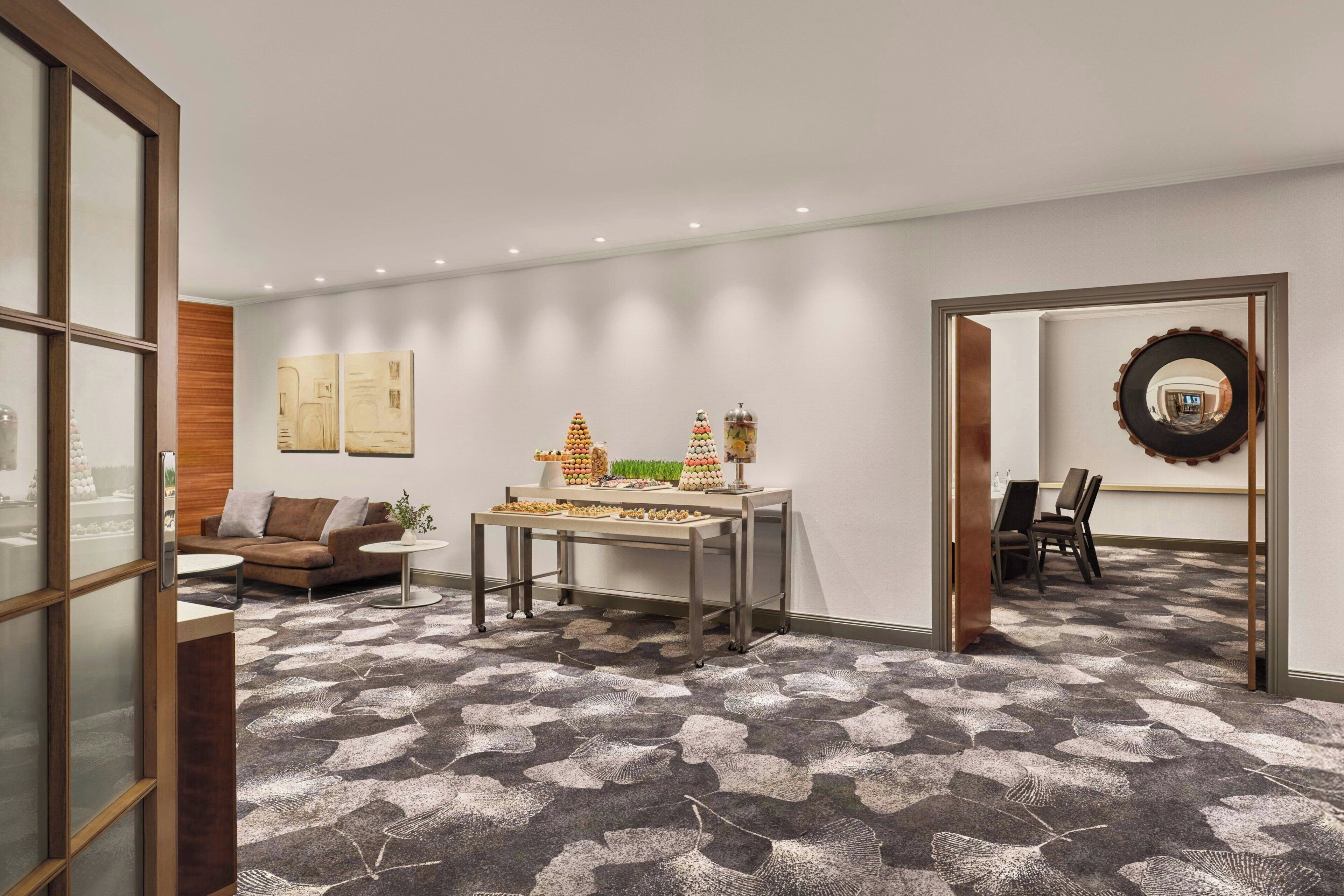 A modern hotel room with a patterned carpet, a dining table with a centerpiece, a sofa, and a mirror on the wall.