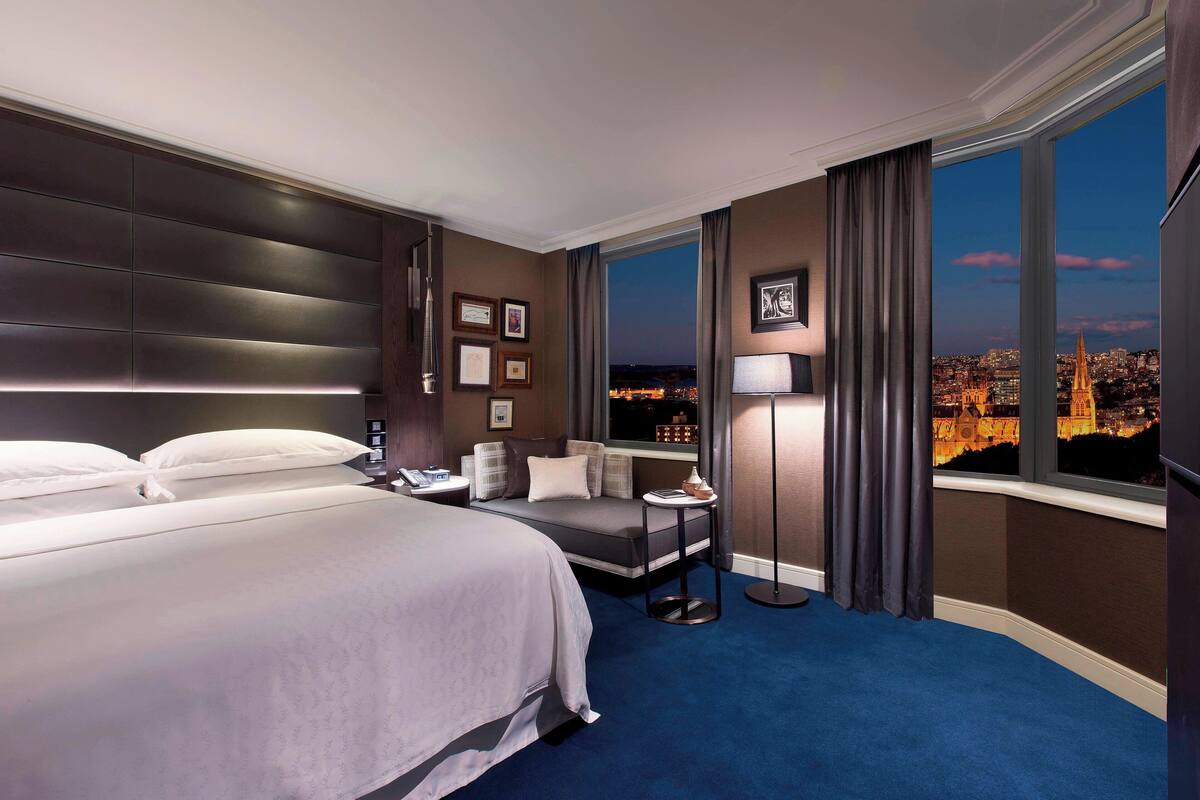 Een hotelkamer met een groot bed, een zithoek, een nachtkastje, een lamp en uitzicht op een stadsgezicht door een groot raam.