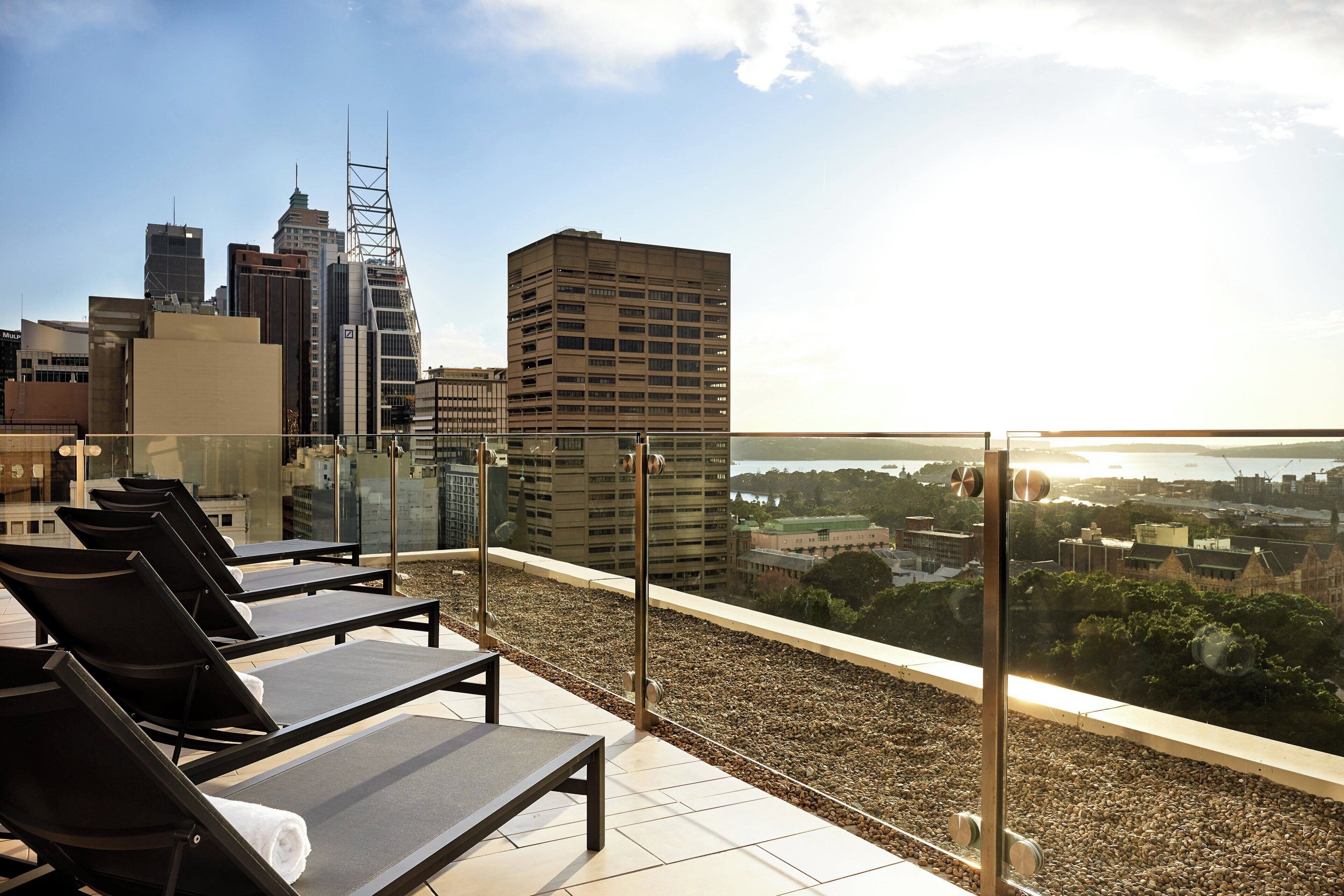 A rooftop terrace with sun loungers, a city skyline view, and a glass railing.