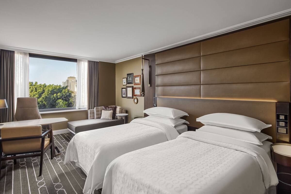 Een hotelkamer met twee bedden, een bureau, een stoel, een raam met gordijnen en uitzicht op de stad.