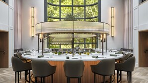 A round dining table with six chairs, surrounded by a wooden panel wall and large windows.