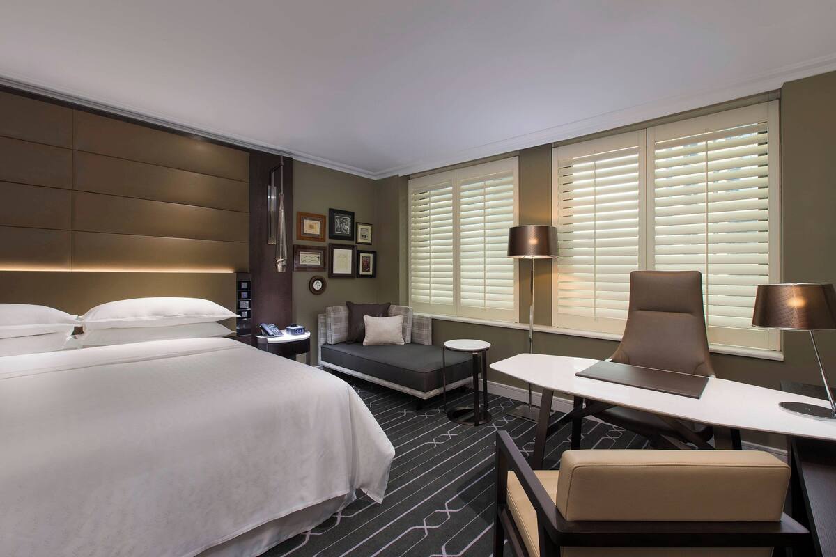 Een hotelkamer met een groot bed, een slaapbank, een bureau en een stoel. Twee nachtkastjes, een lamp en een raam met jaloezieën zijn aanwezig.