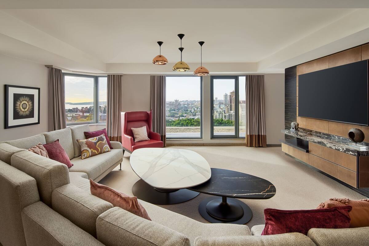 Een moderne woonkamer met een bank, fauteuil en een rond salontafel. Er is een grote televisie aan de muur en er is uitzicht op de stad door de ramen.