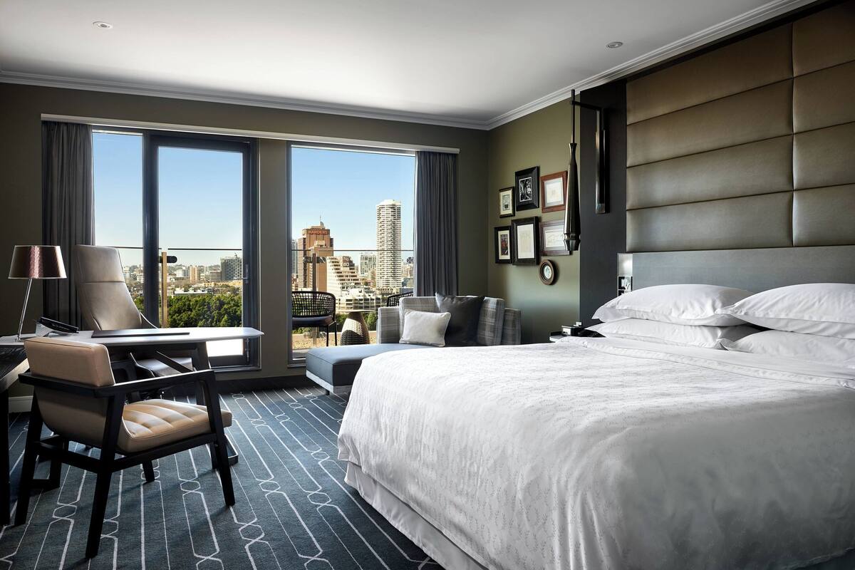 Een hotelkamer met een groot bed, een bureau en een stoel. Grote ramen bieden uitzicht op de stad.