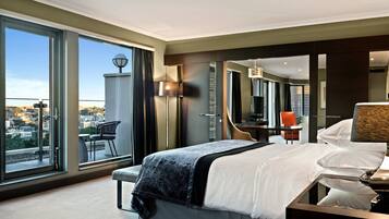 A modern hotel room with a large bed, a sitting area with a chair, a desk, and a large window offering a city view.