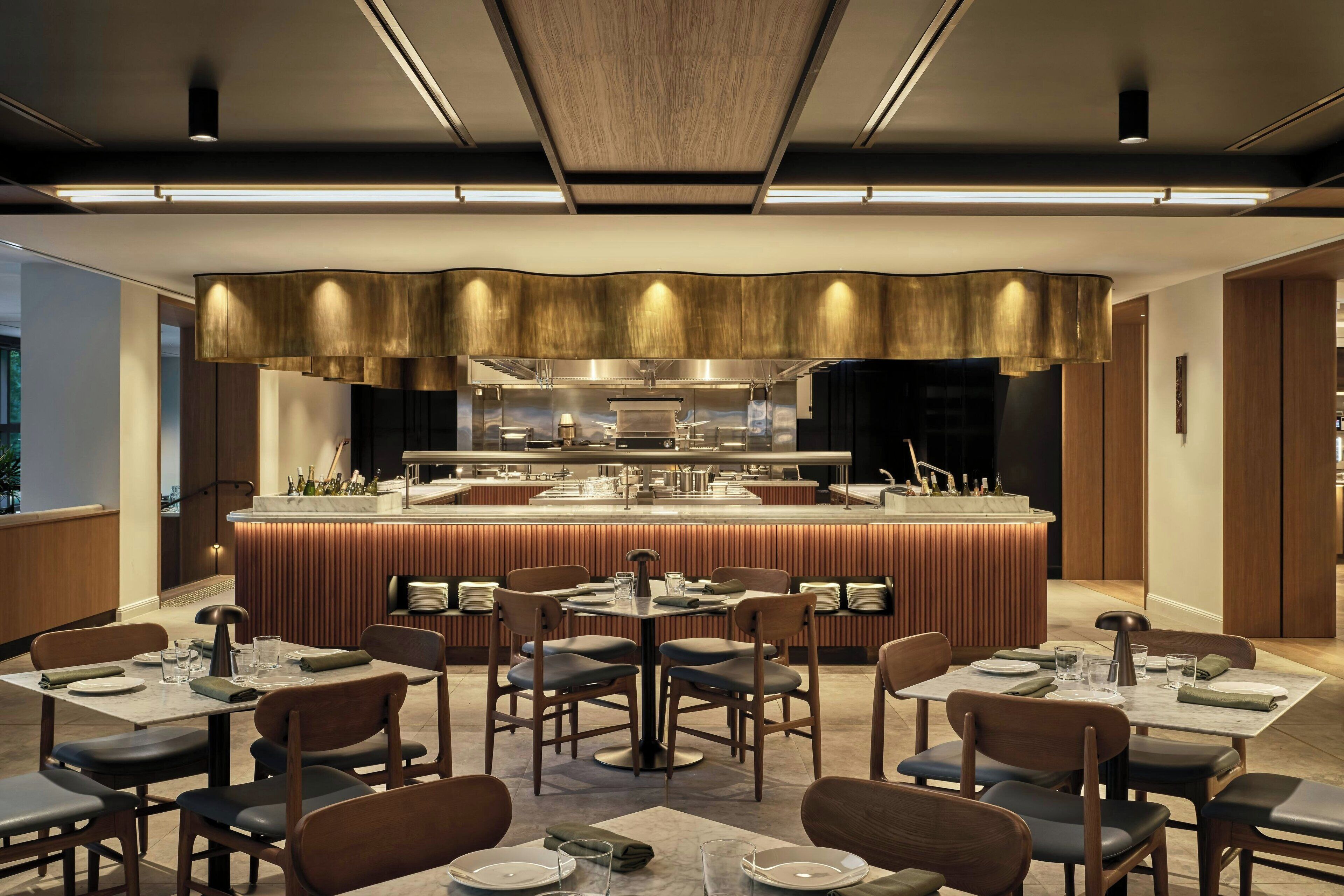 A modern restaurant with a central bar, multiple dining tables, and a contemporary kitchen in the background.