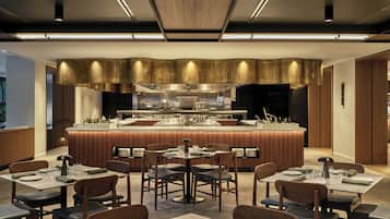 A modern restaurant with a central bar, multiple dining tables, and a contemporary kitchen in the background.