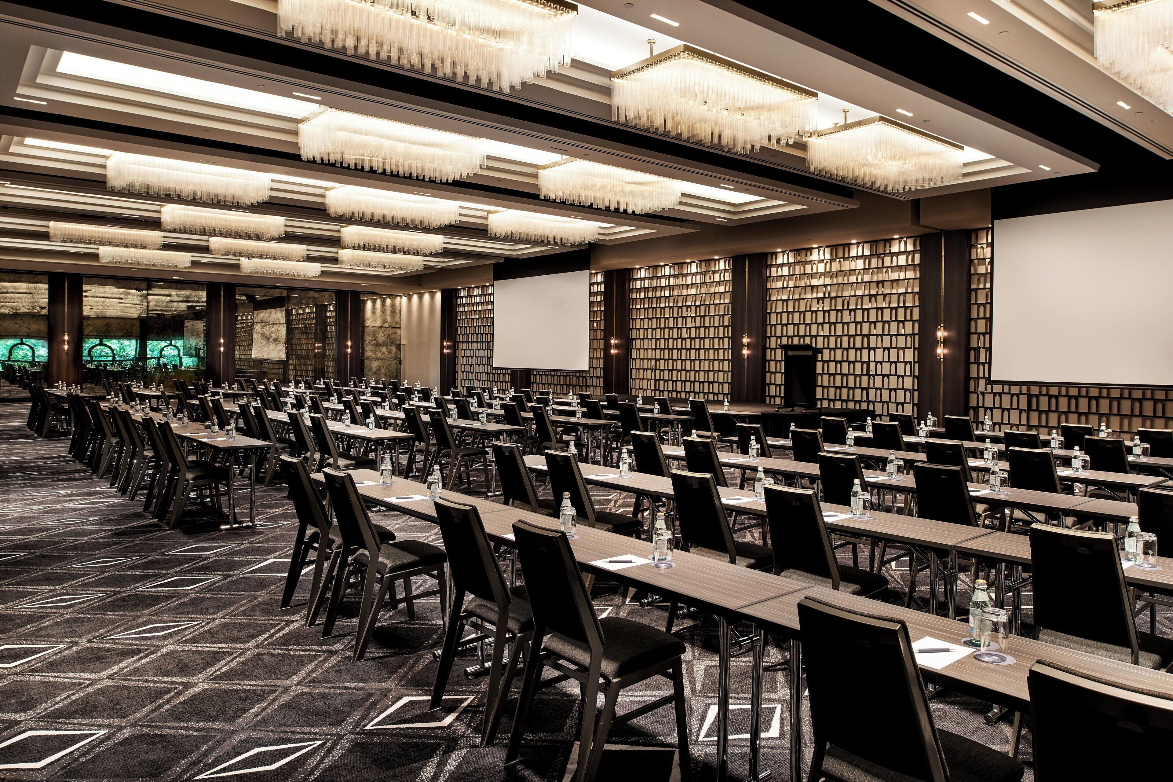 Une grande salle de conférence avec de longues rangées de tables et de chaises, des lustres et un écran de projection.