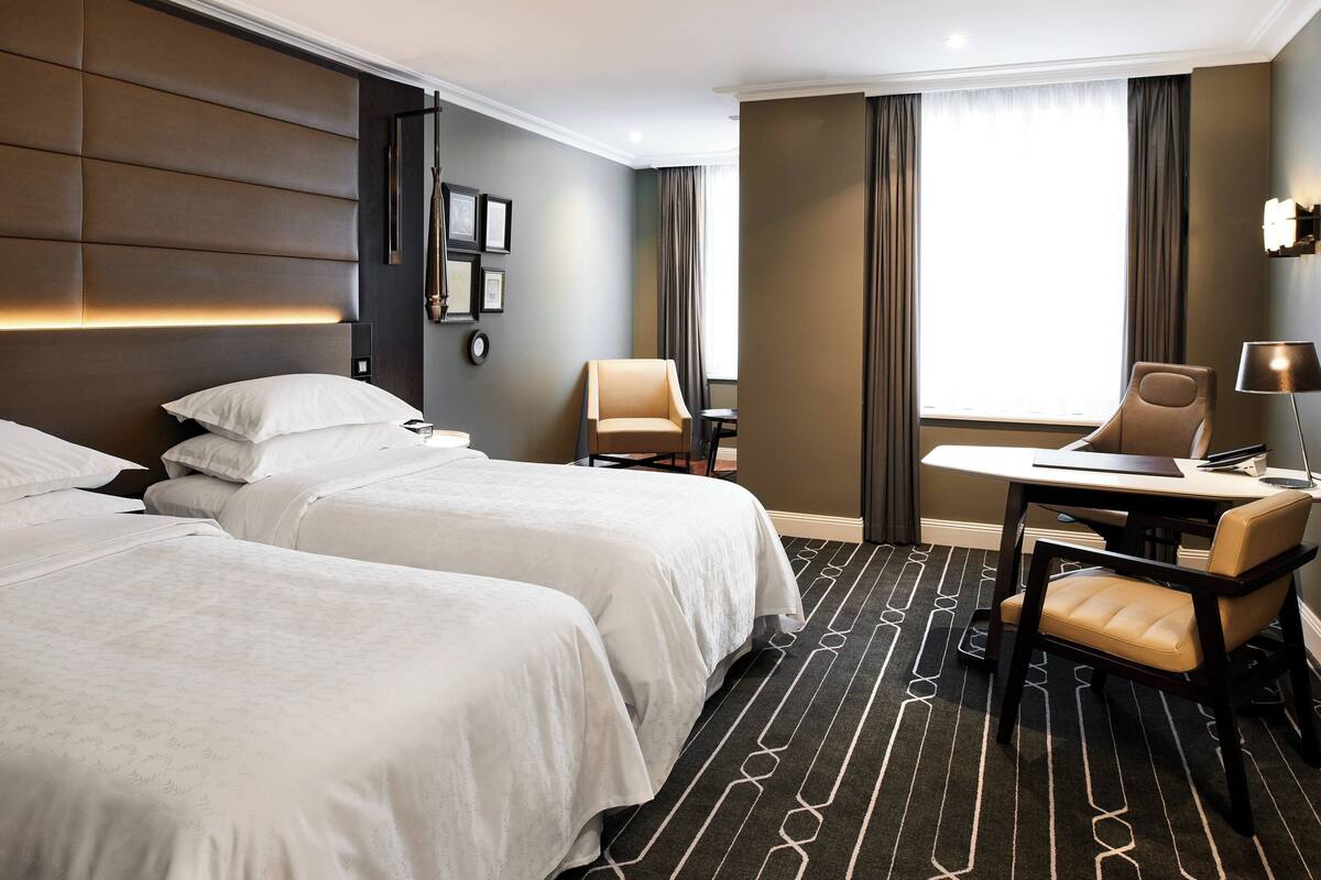 Een hotelkamer met twee bedden, een bureau met stoel, een lamp en een groot raam met gordijnen.