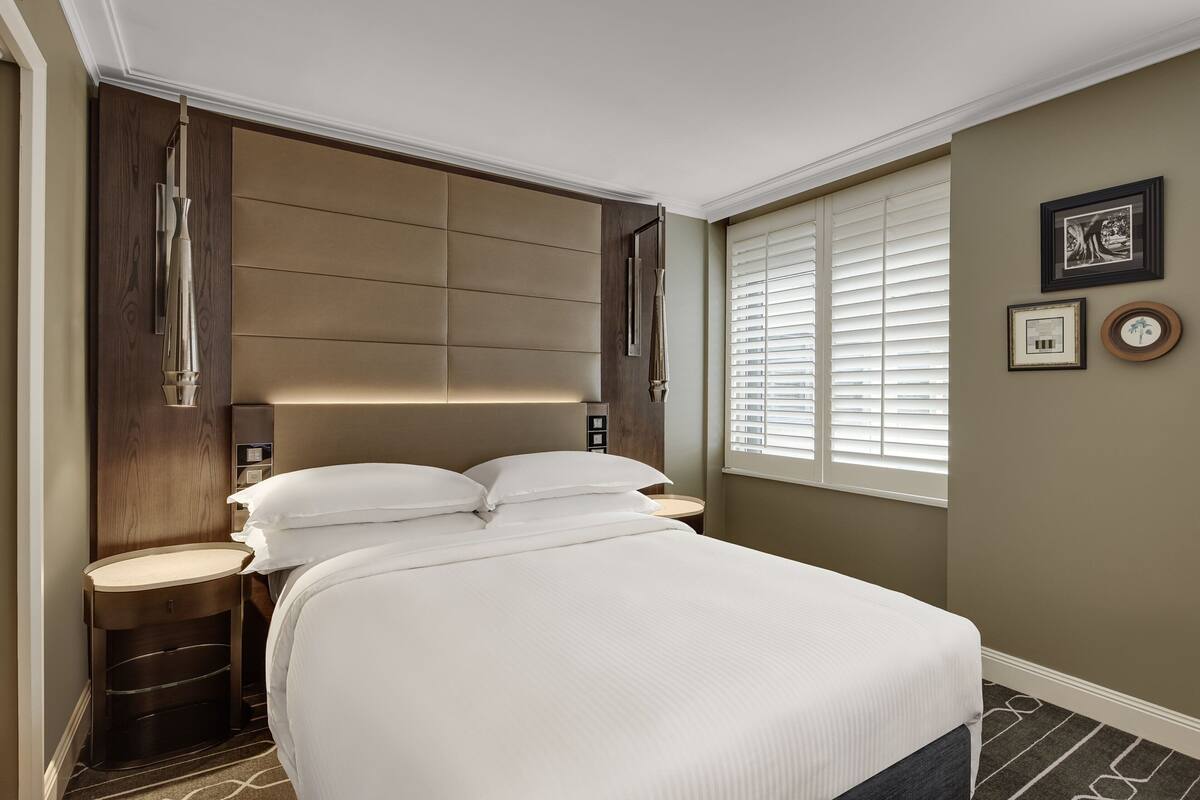 Een hotelkamer met een groot bed, een houten hoofdbord, twee nachtkastjes en een raam met jaloezieën.