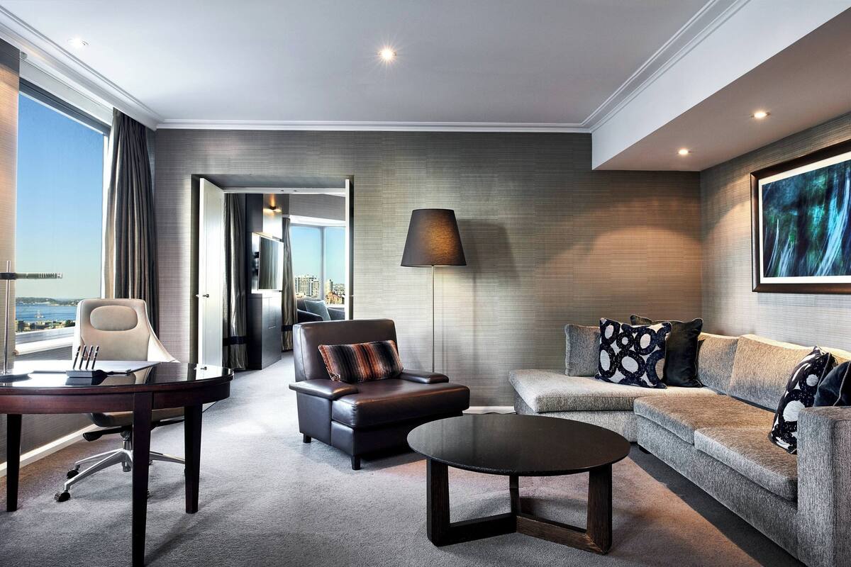 Een moderne hotelkamer met een leren bank, een ronde salontafel, een bureau met stoel, en een groot raam met uitzicht op de stad.