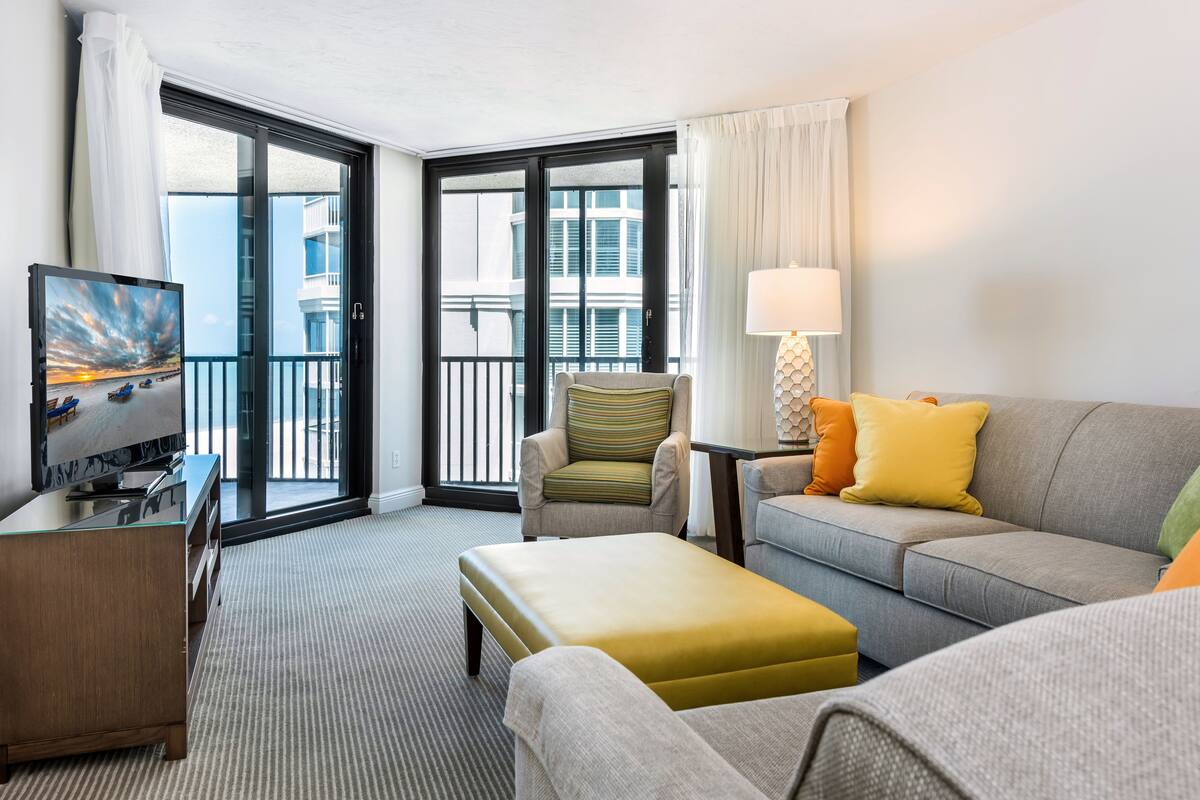 A hotel room with a sofa, armchair, ottoman, TV, and a view of the city through the balcony doors.