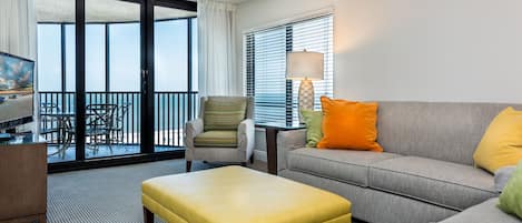 A hotel room with a balcony, a sofa, an ottoman, a chair, a lamp, and a TV.