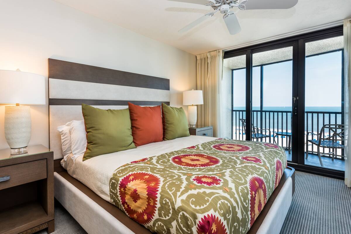 A hotel room with a large bed, a ceiling fan, a balcony with chairs, and a view of the ocean.