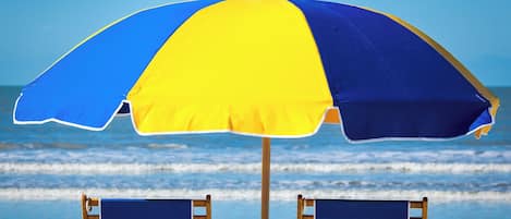 A beach umbrella with a blue and yellow design, two wooden beach chairs, and a sandy beach with waves in the background.