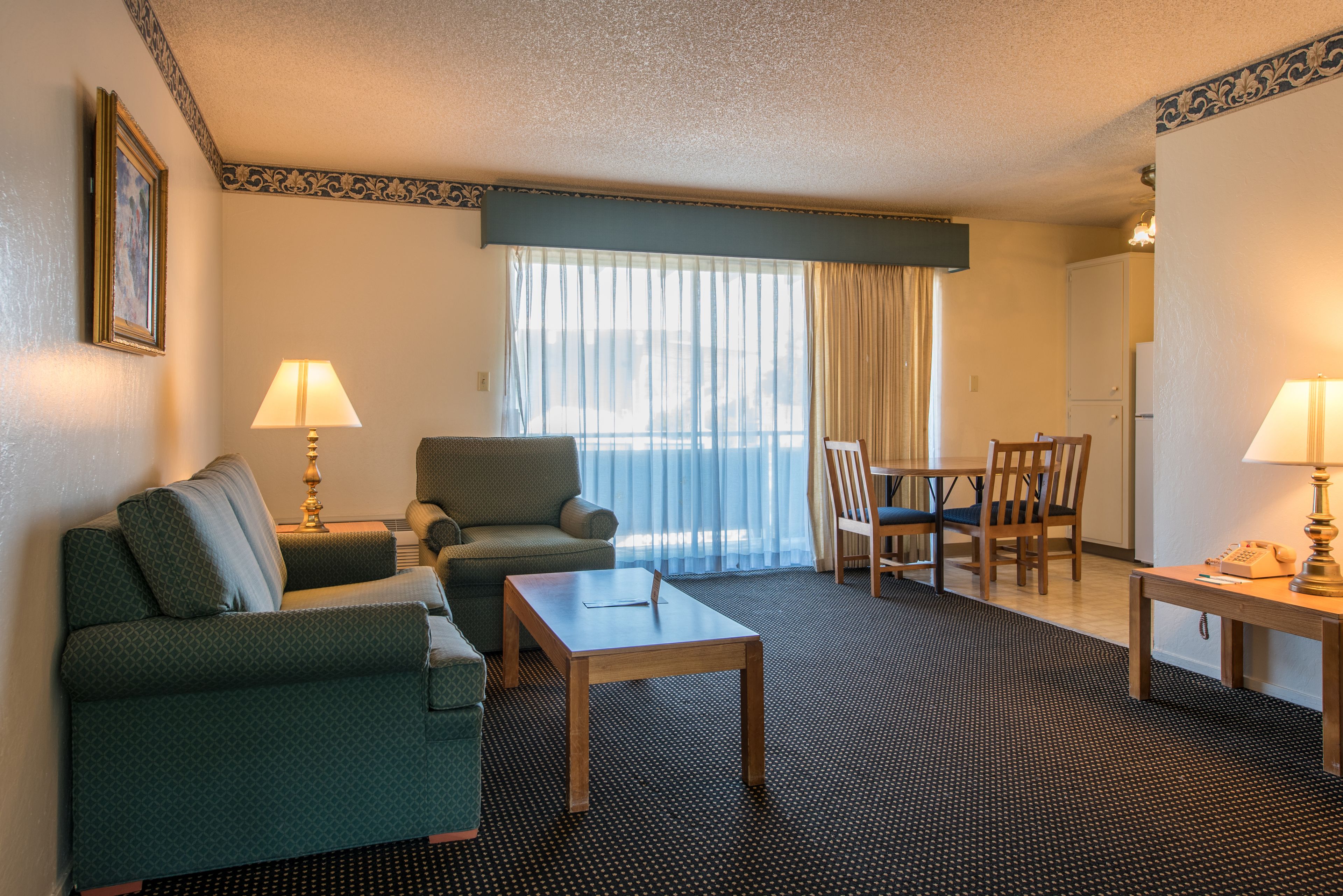 Habitación de hotel con un sofá verde, una mesa de centro de madera, una mesa de comedor con sillas y un ventanal con cortinas.