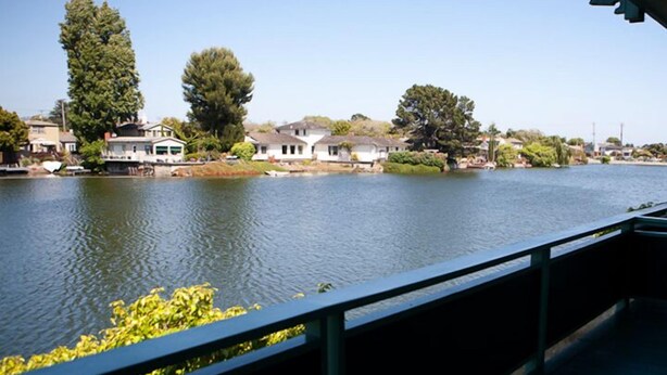 Une rivière bordée de maisons sur la rive opposée, un ciel dégagé et une rambarde de pont au premier plan.