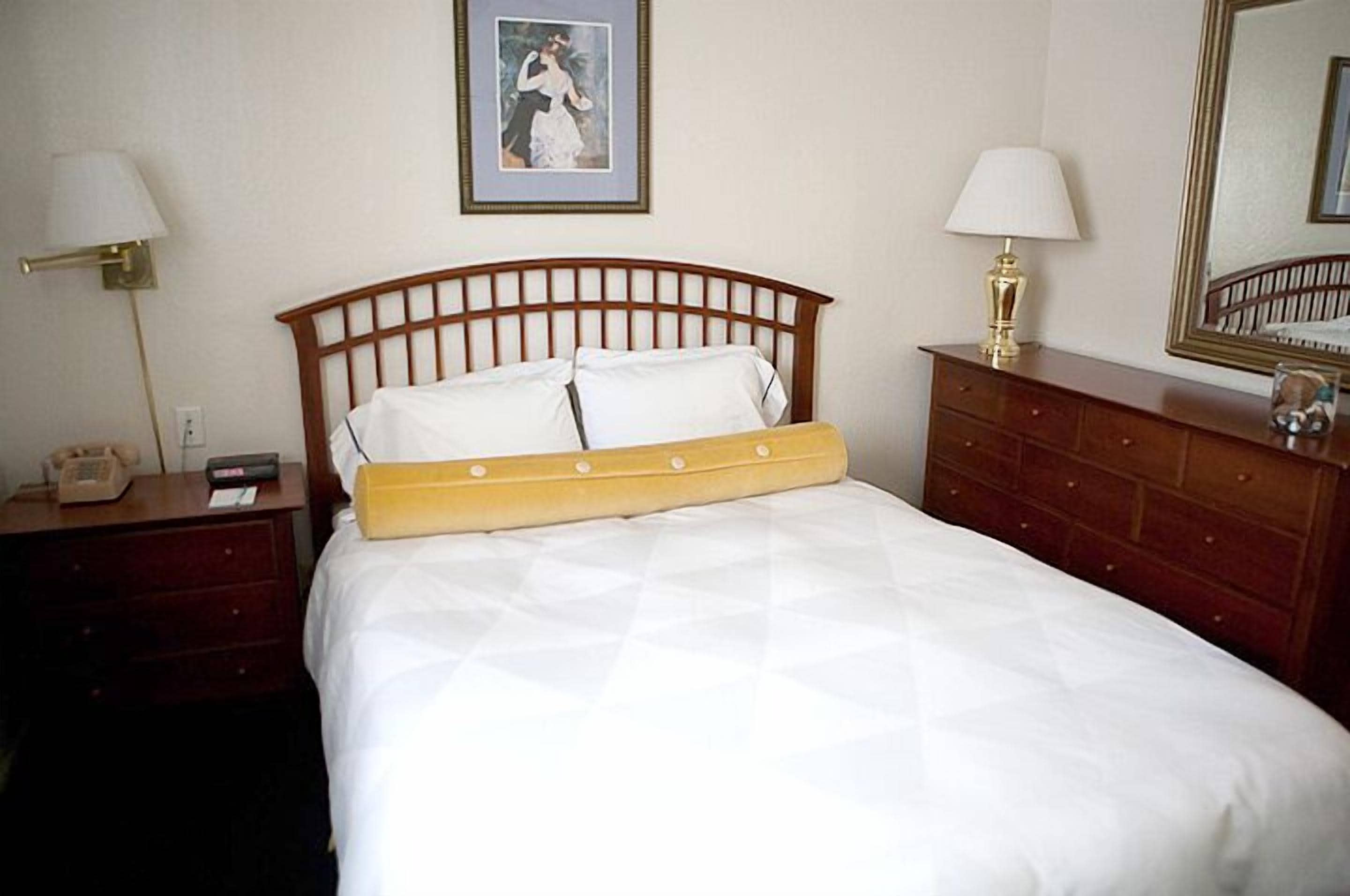 Een keurig opgemaakt bed met houten hoofdbord, een nachtkastje met lamp, een dressoir en een ingelijst foto aan de muur.