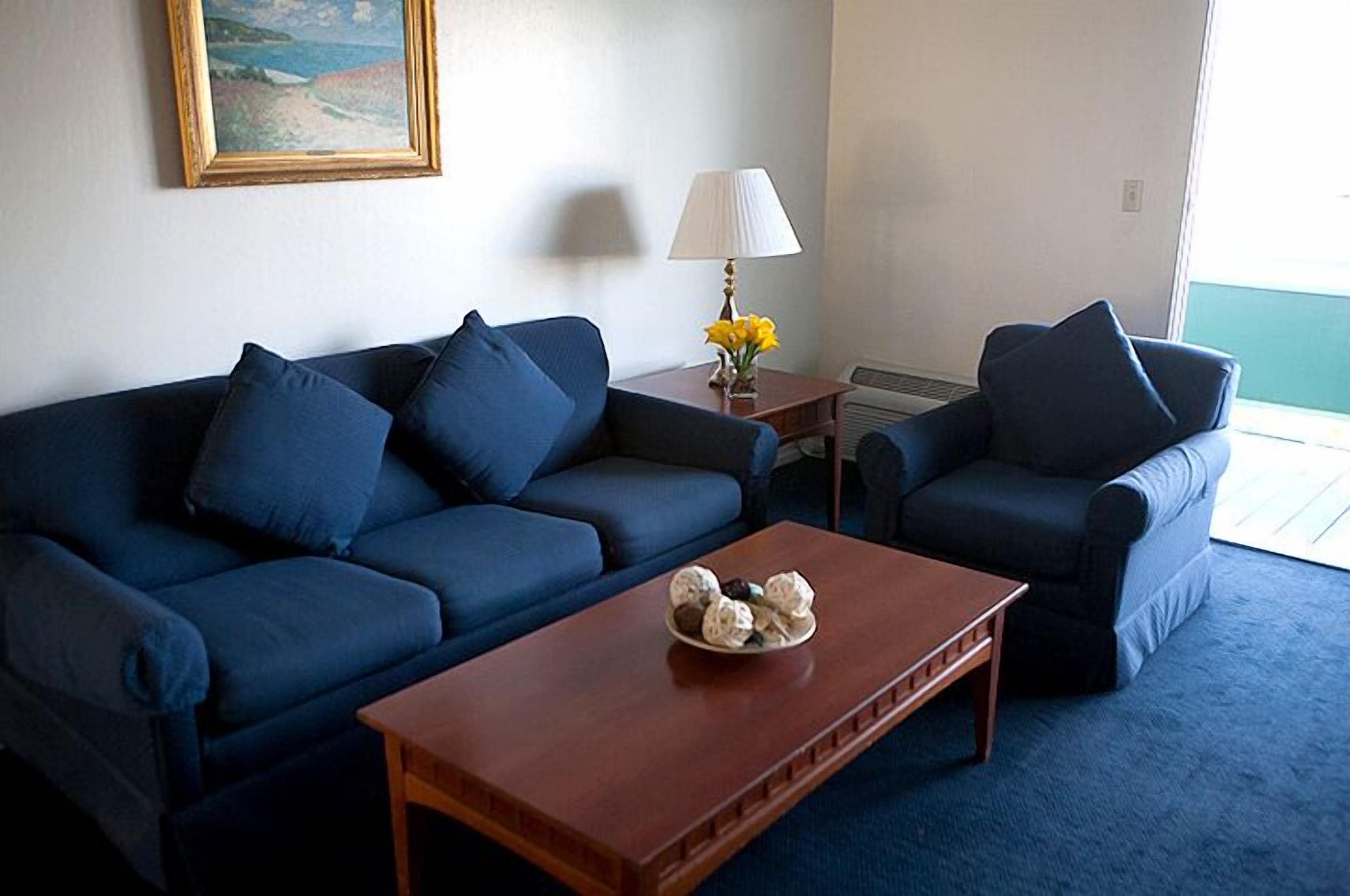 Un salon avec un canapé bleu, un fauteuil assorti, une table basse en bois, une lampe et un tableau accroché au mur.