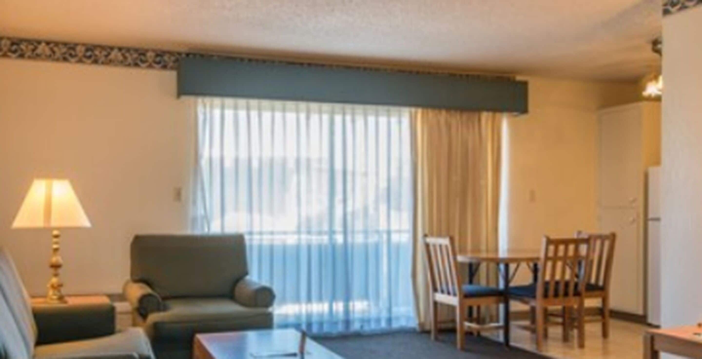Habitación de hotel con sofá, una mesita, una lámpara, una mesa de comedor con sillas y un ventanal con cortinas.