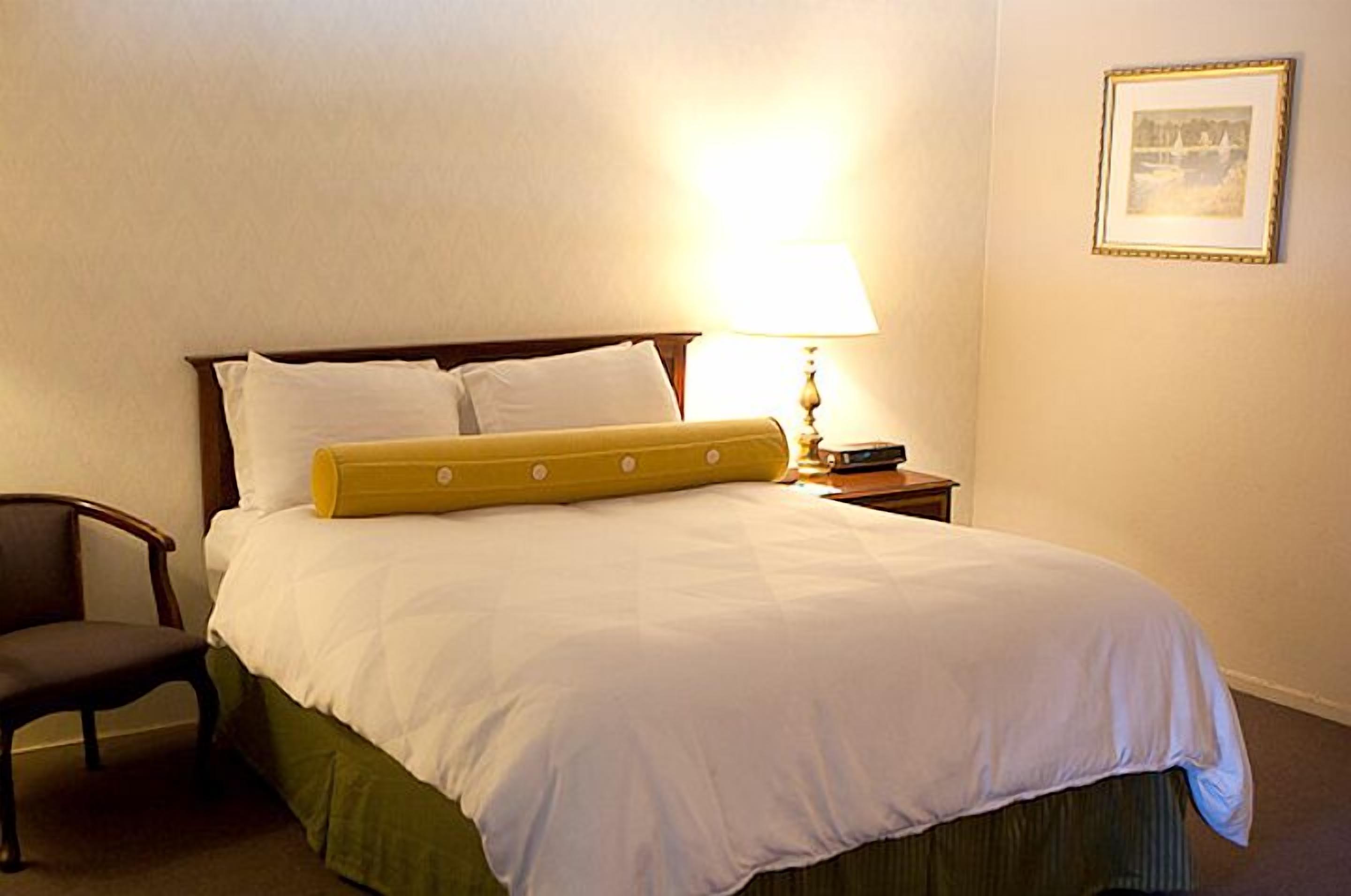 A neatly made bed with white bedding and a yellow accent pillow, a bedside lamp, a framed picture on the wall, and a chair to the left of the bed.