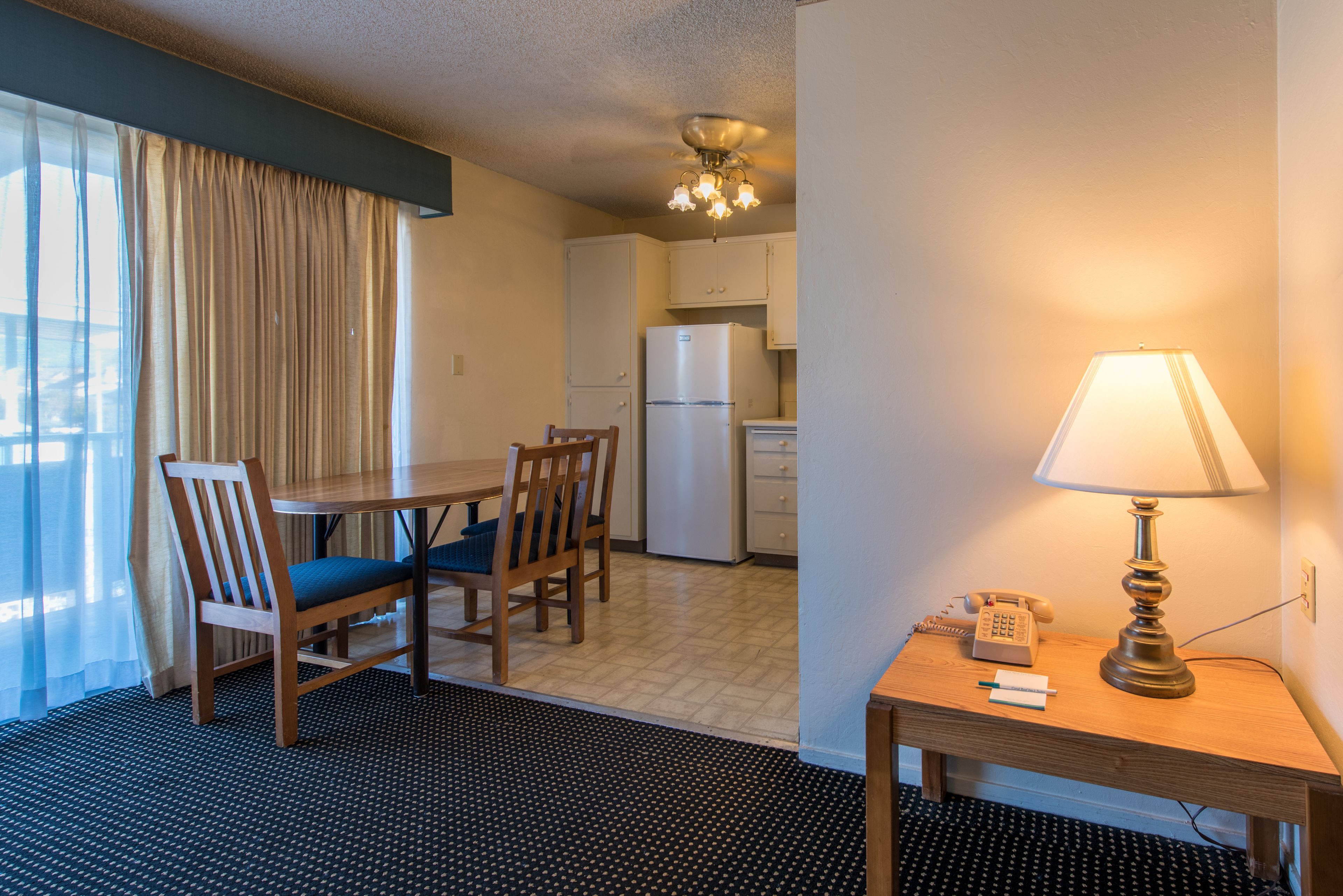 A hotel room with a dining table, chairs, a lamp, a phone, and a refrigerator.