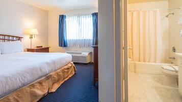 A hotel room with a large bed, a bedside table with a lamp, a window with curtains, an air conditioning unit, and a bathroom with a bathtub and toilet.