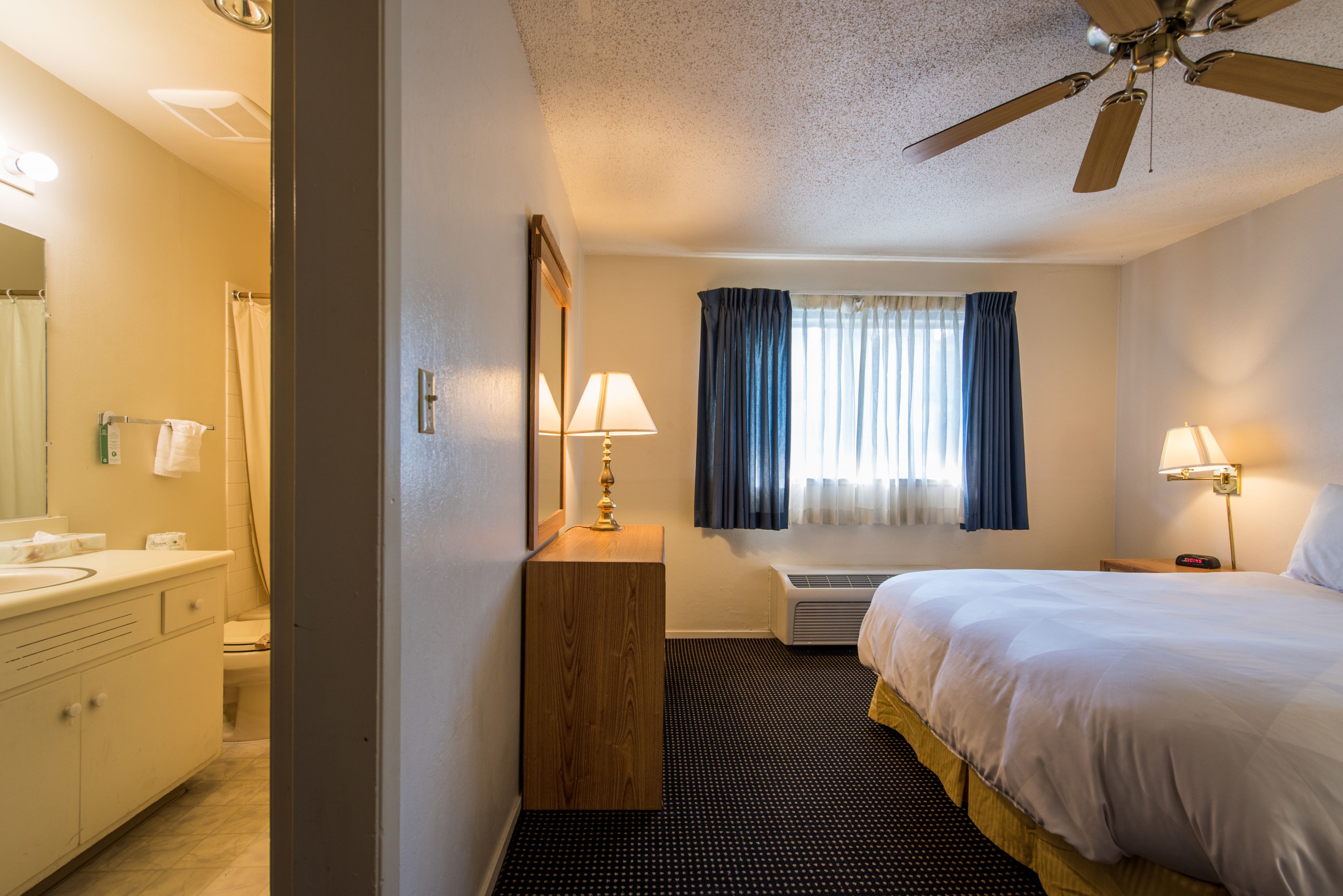 A hotel room with a bed, bedside lamps, a ceiling fan, a window with curtains, a bathroom with a sink and mirror, and a small wooden nightstand.