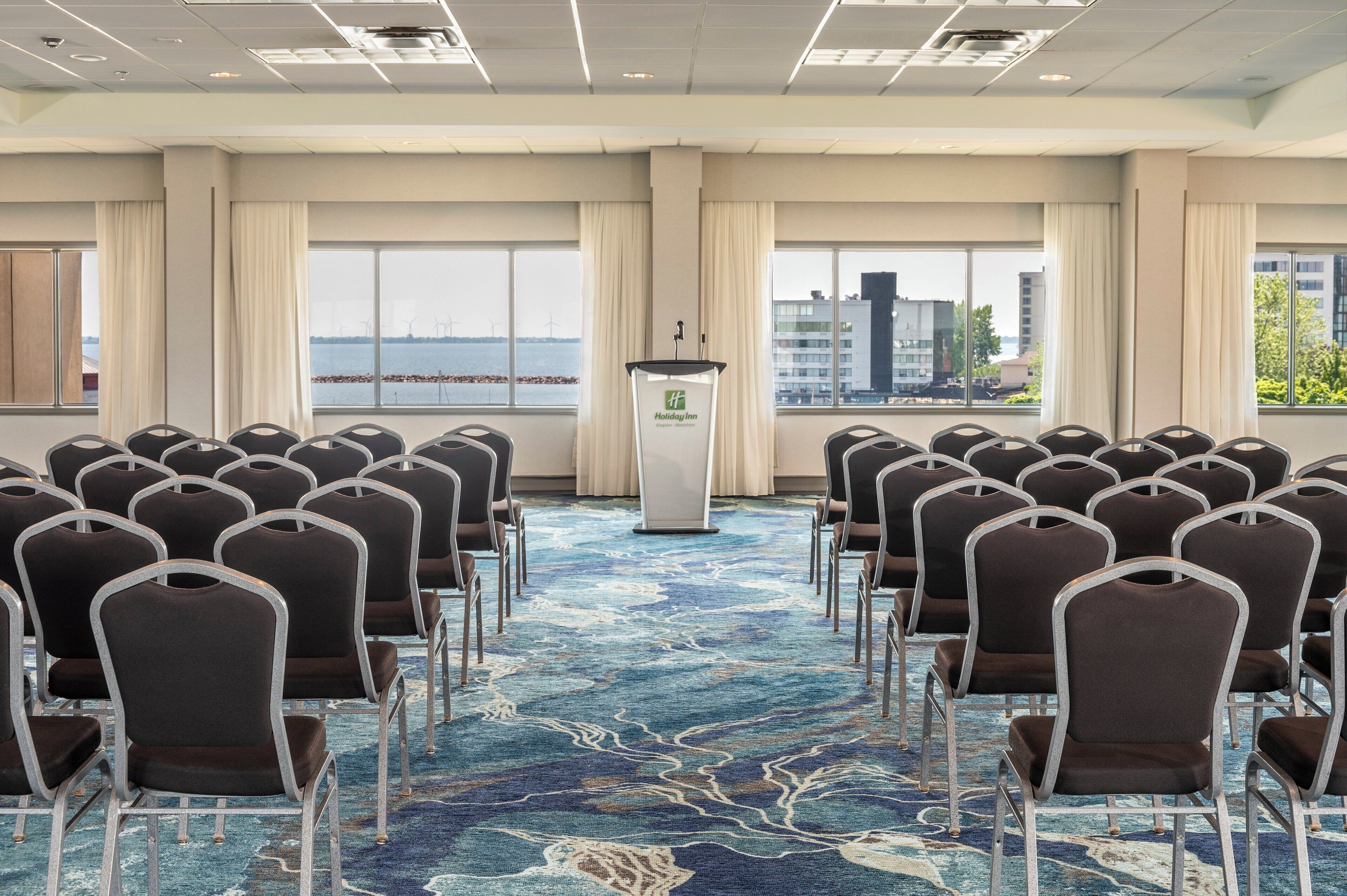 A conference room with rows of chairs facing a podium, large windows with a view of buildings and a body of water, and a carpet with a blue and white pattern.