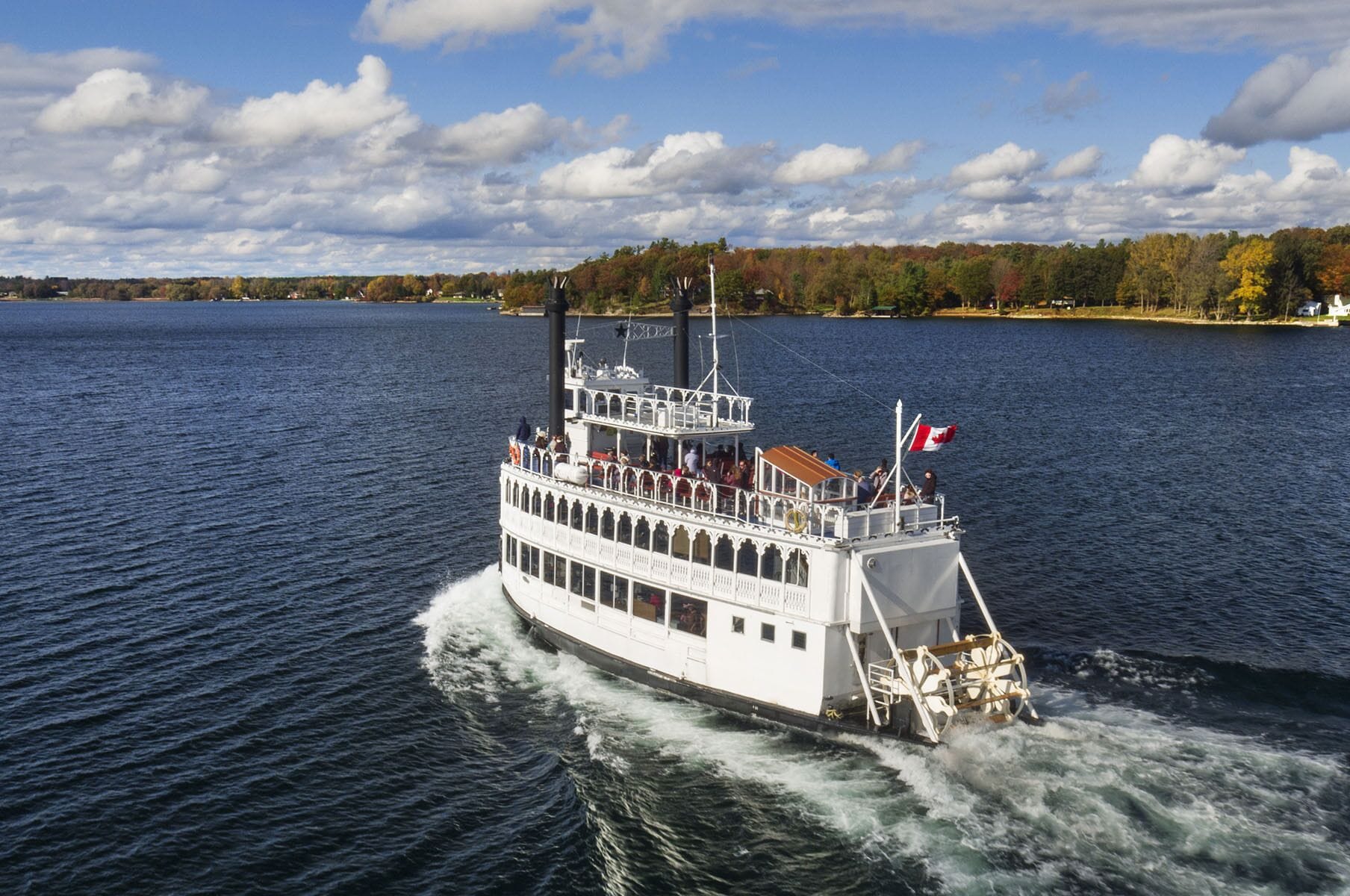 A white river cruise ship with a Canadian flag, multiple decks, and passengers enjoying the view.