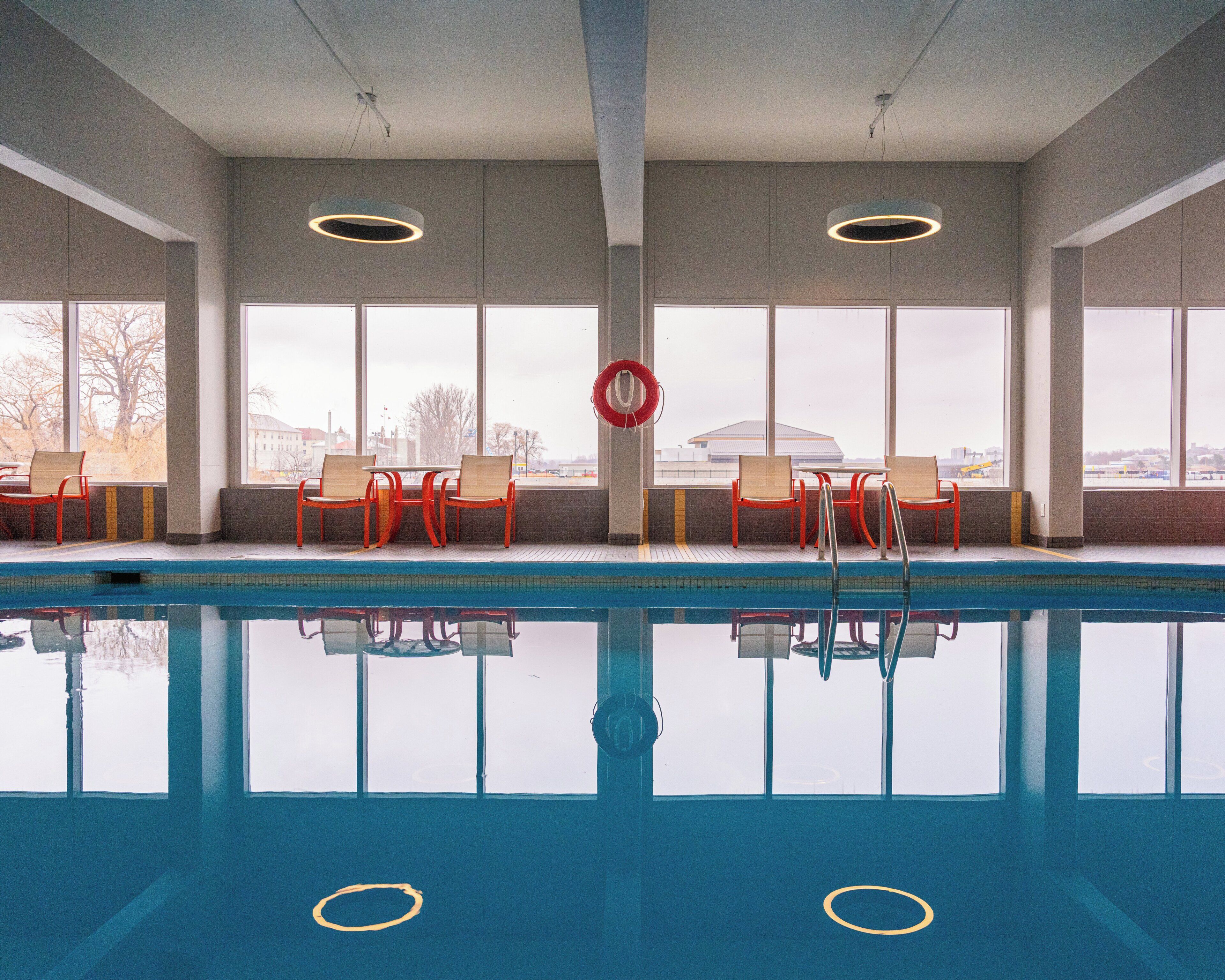 Una piscina interior con agua cristalina, sillas rojas de madera y grandes ventanales con vistas a la ciudad.