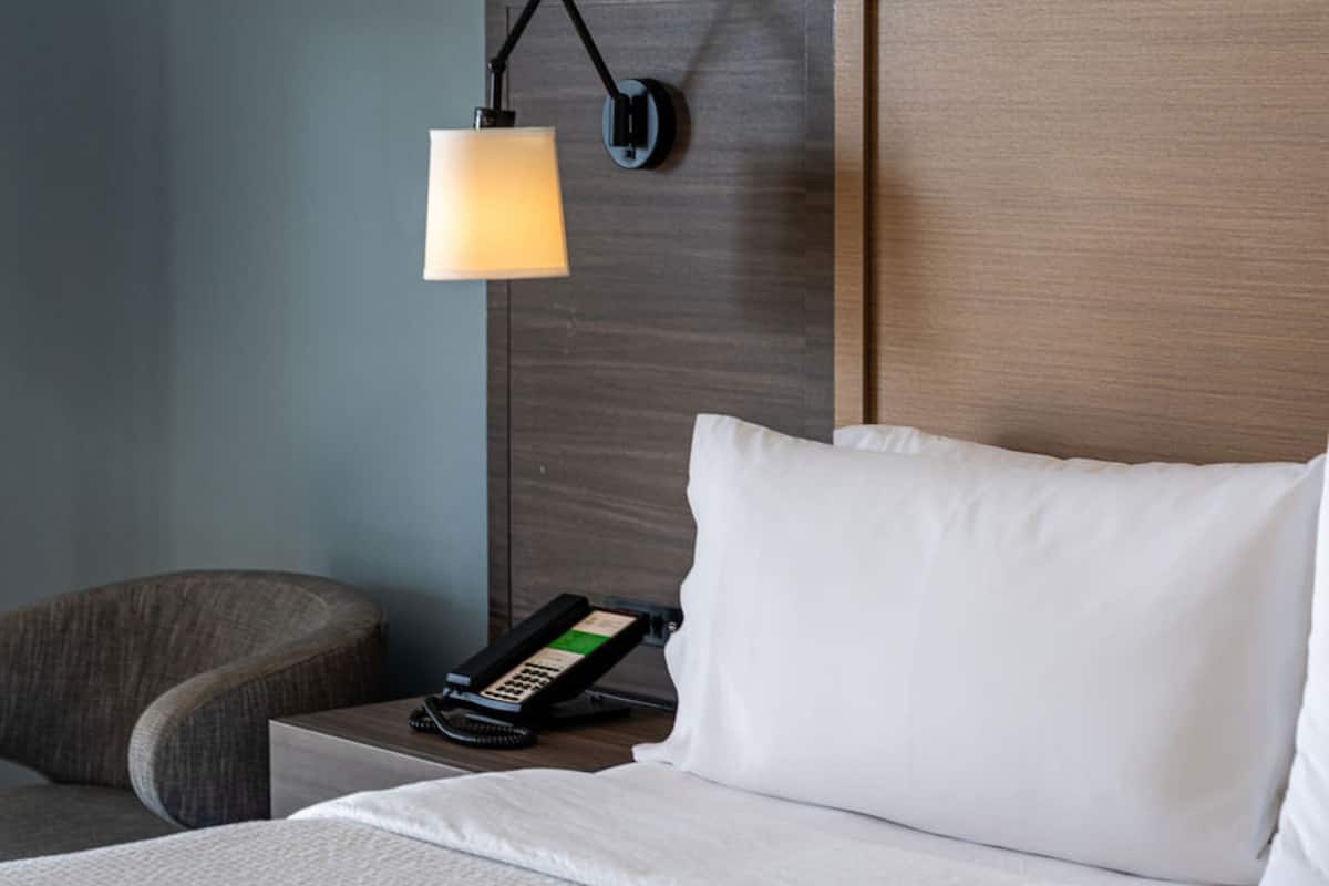 A hotel room with a bed, a bedside phone, a lamp, and a chair.