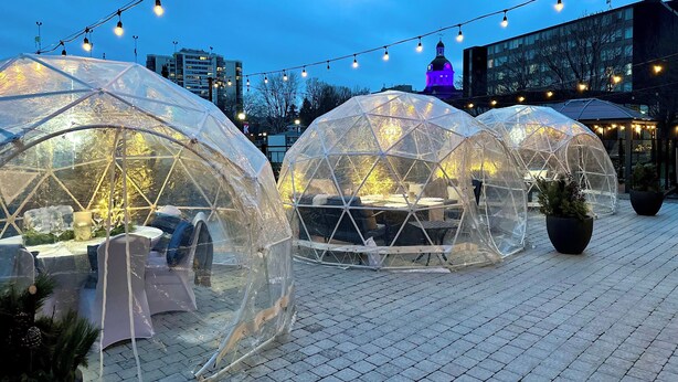 Des dômes géodésiques transparents, aménagés avec des sièges à l’intérieur, reliés par des guirlandes lumineuses, et installés sur une surface pavée.