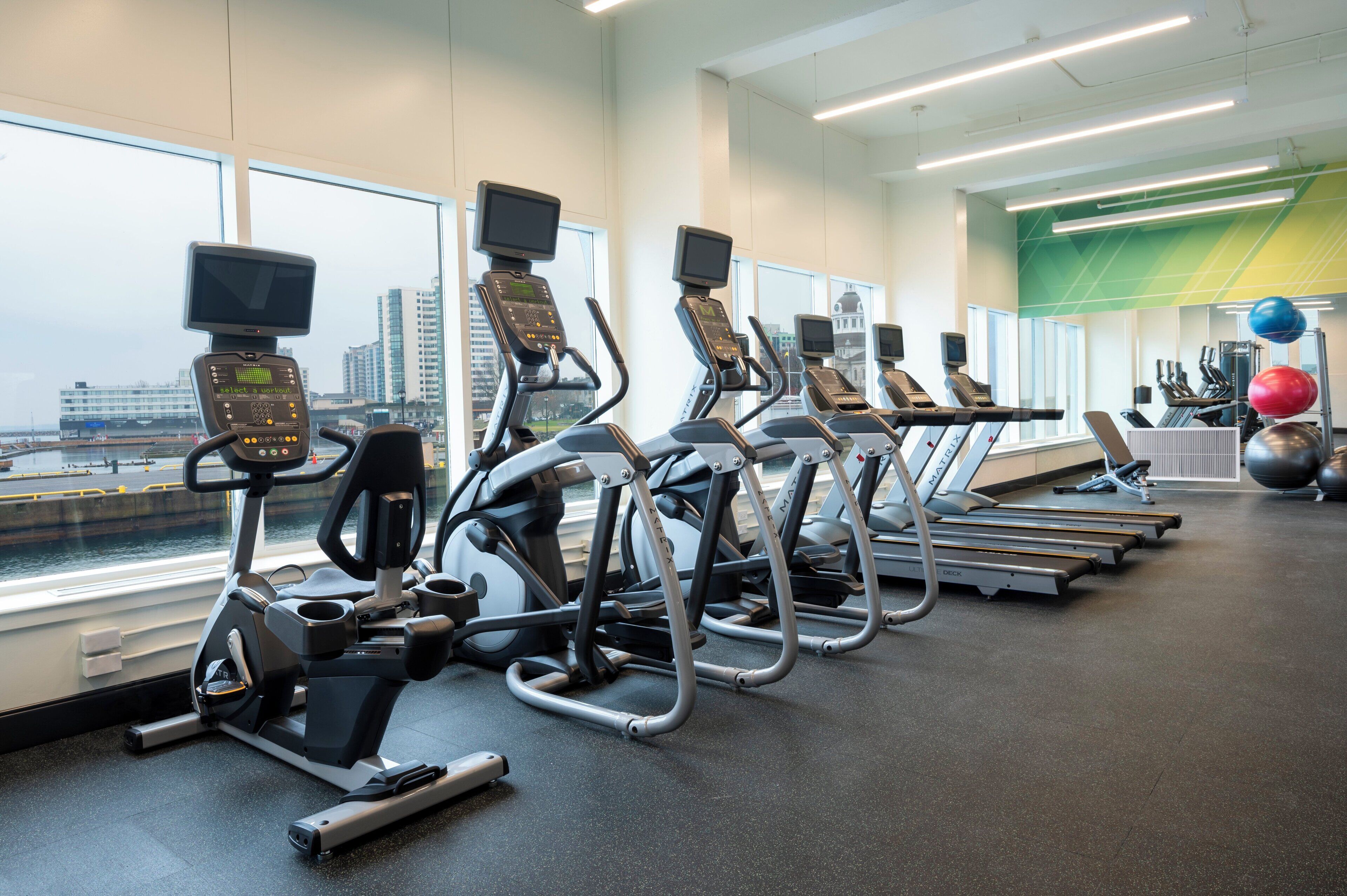 A gym with treadmills, elliptical machines, and exercise balls, featuring large windows with a city view.