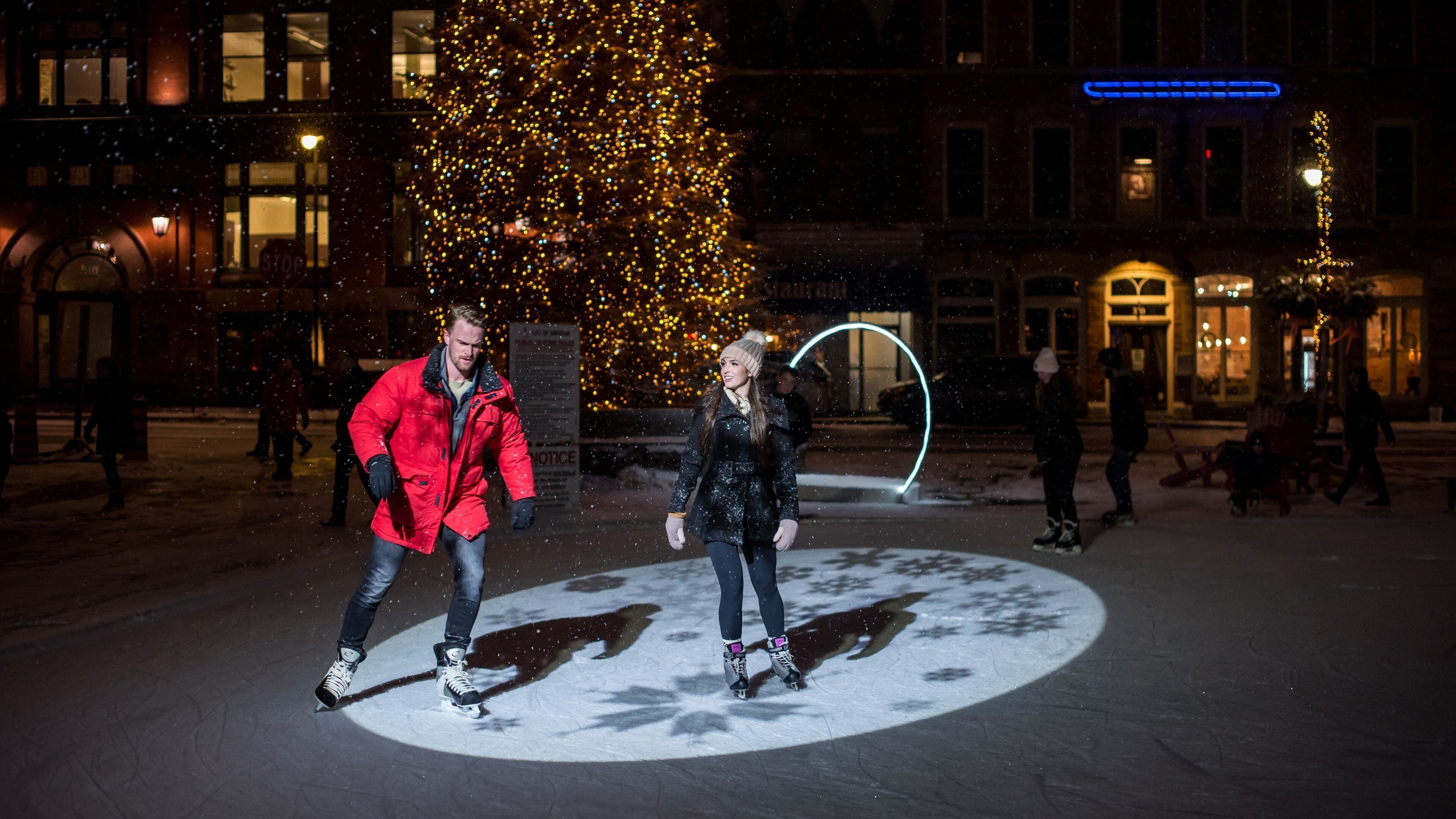 A nighttime outdoor ice skating area with a large illuminated Christmas tree and a neon sign in the background.
