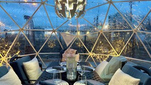 Une structure en forme de dôme géodésique abrite une table à manger pour quatre personnes, agrémentée de sets de table tissés, de verres et d’un centre de table décoratif.