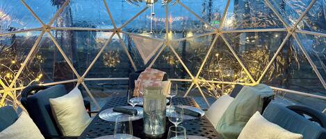 A geodesic dome structure with a dining table set for four, featuring woven placemats, glasses, and a decorative centerpiece.