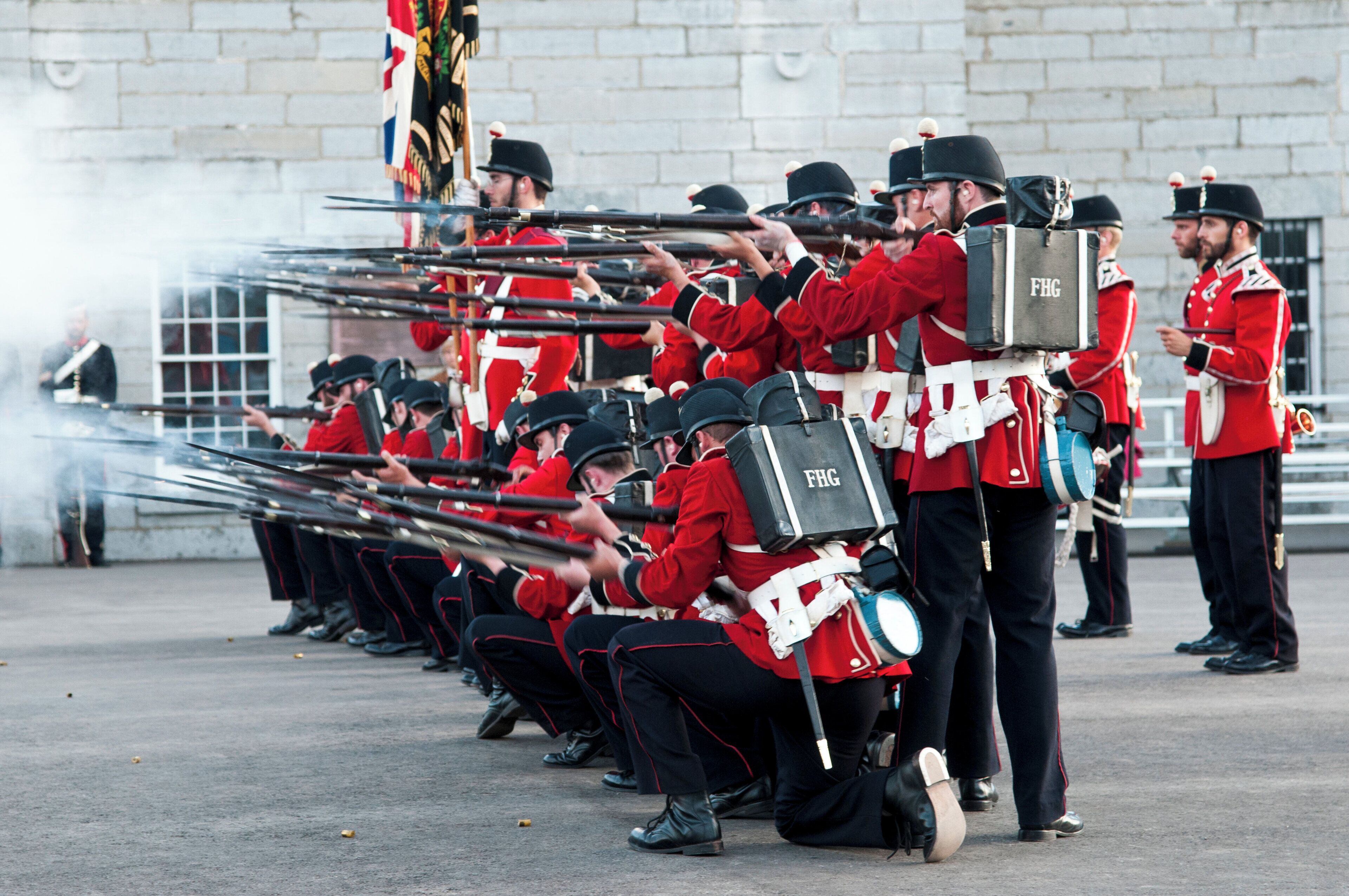 A group of uniformed personnel in red and black attire, carrying rifles and a large bag marked "FHG".