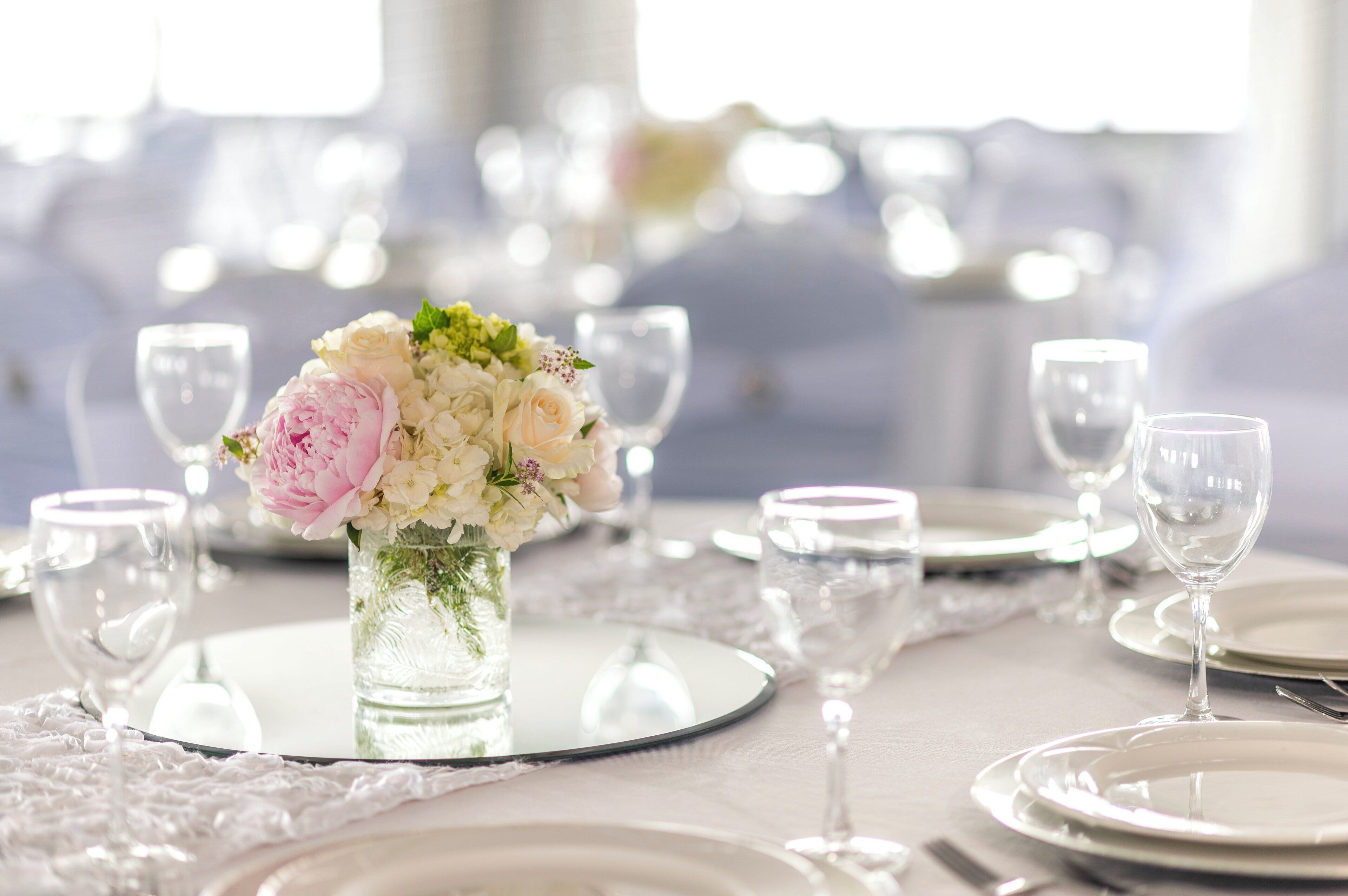 Une table dressée avec de l’assiette blanche, des verres à vin transparents et un vase contenant des fleurs blanches et roses.